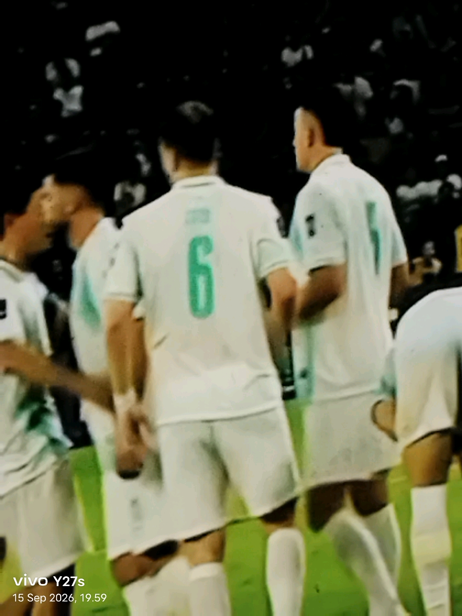 The image shows several individuals in white sports uniforms with green accents. One individual in the foreground has the number "6" prominently displayed on the back of their jersey in green. The other individuals are partially visible, also wearing similar uniforms. They appear to be on a grassy field, likely a sports venue, with a dark background, possibly spectators or stadium lights. The bottom left corner of the image contains a watermark that reads "vivo Y27s" and "15 Sep 2026, 19.59." The image quality is low, with significant blur and pixelation, making it difficult to discern fine details. There is no discernible information within the image that would allow for the identification of the specific location in Tamansari, Indonesia, or any specific actions or interactions occurring.