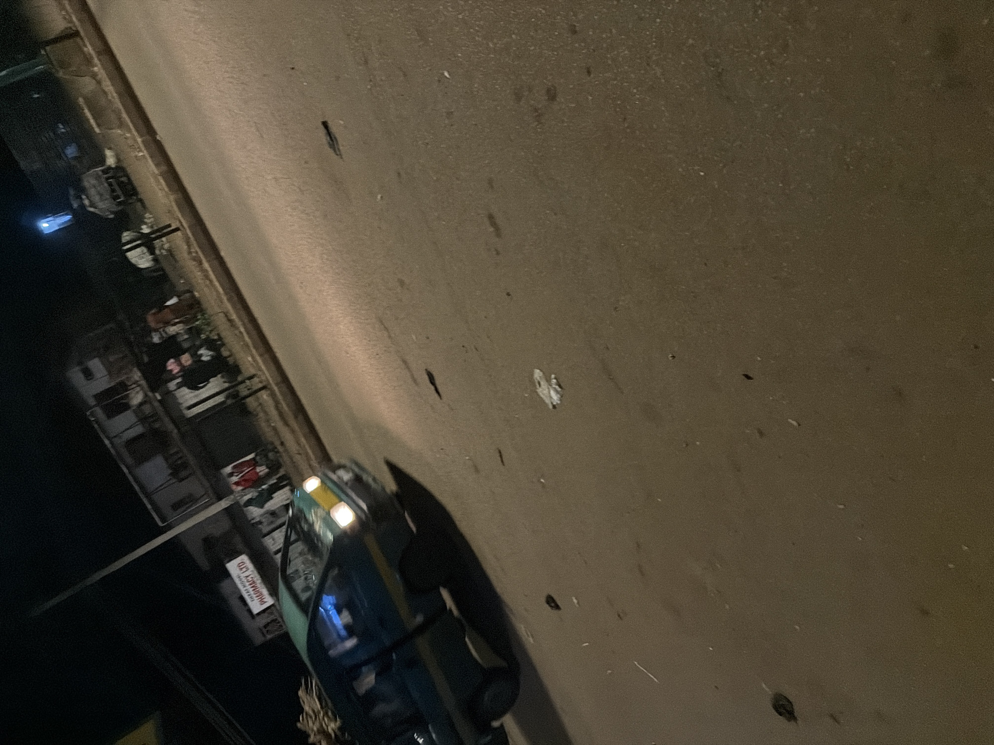 The image shows a street scene at night in Bukuru, Nigeria. A teal-colored van with yellow trim and its headlights on is visible in the foreground, moving from left to right. The van appears to be carrying goods on its roof. In the background, a building with a sign that reads "PHARMACY LTD" is visible. Several individuals are standing near the building. The street is paved and appears to be illuminated by streetlights. There is some litter on the ground. The overall impression is that of a busy urban environment at night.