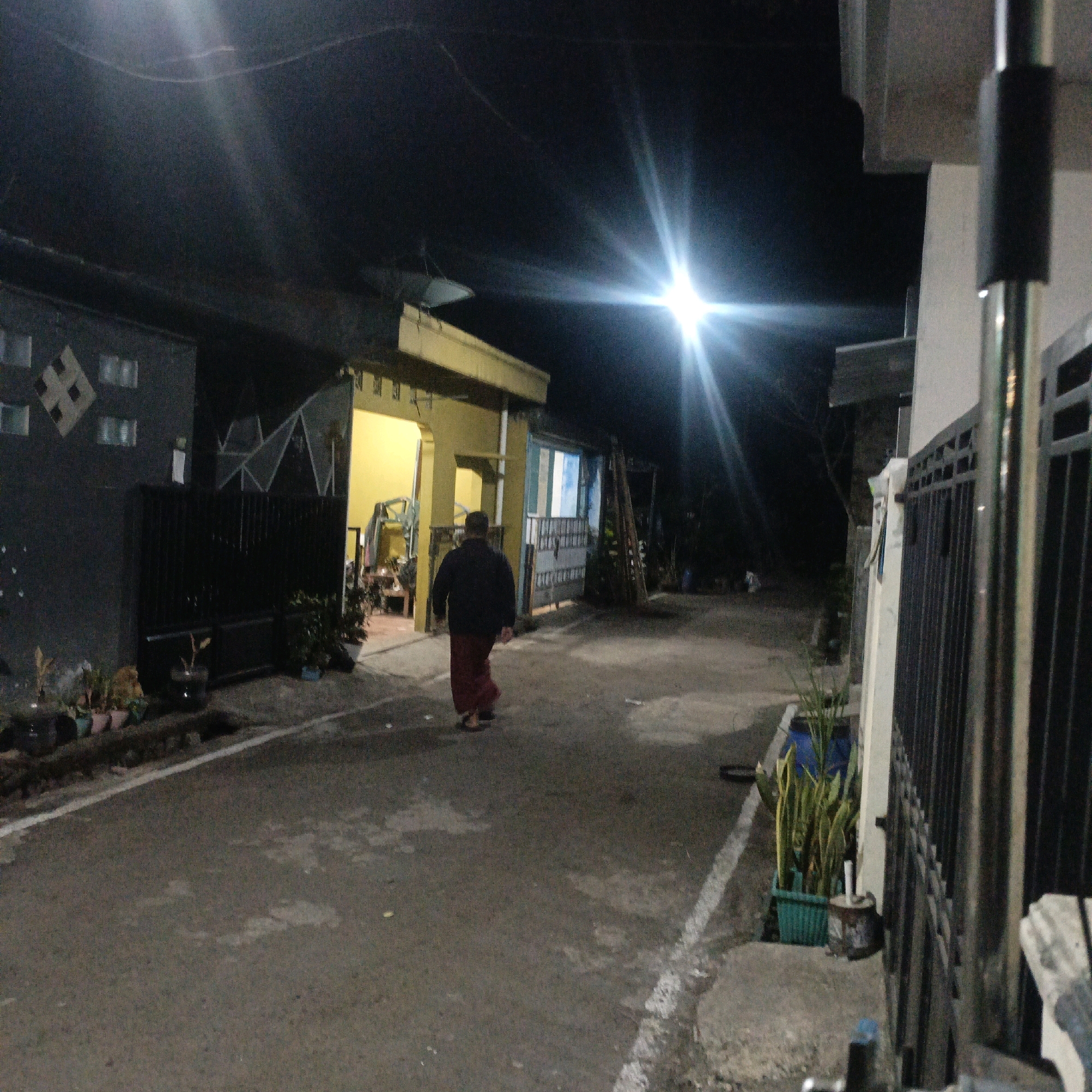 The image depicts a street scene at night in Salatiga, Indonesia, illuminated by artificial lights creating lens flares. A person wearing a dark jacket and a maroon sarong walks away from the camera down a paved street. On the left, a dark-colored building with square window patterns and a black metal fence is visible. In the center, a building with a bright yellow facade has an open doorway revealing interior structures. To the right, a black metal fence runs along the side of the street, and a tall metal pole stands in the foreground. Several potted plants are arranged along the left side of the street near the fence. The overall environment appears to be a residential or commercial area.