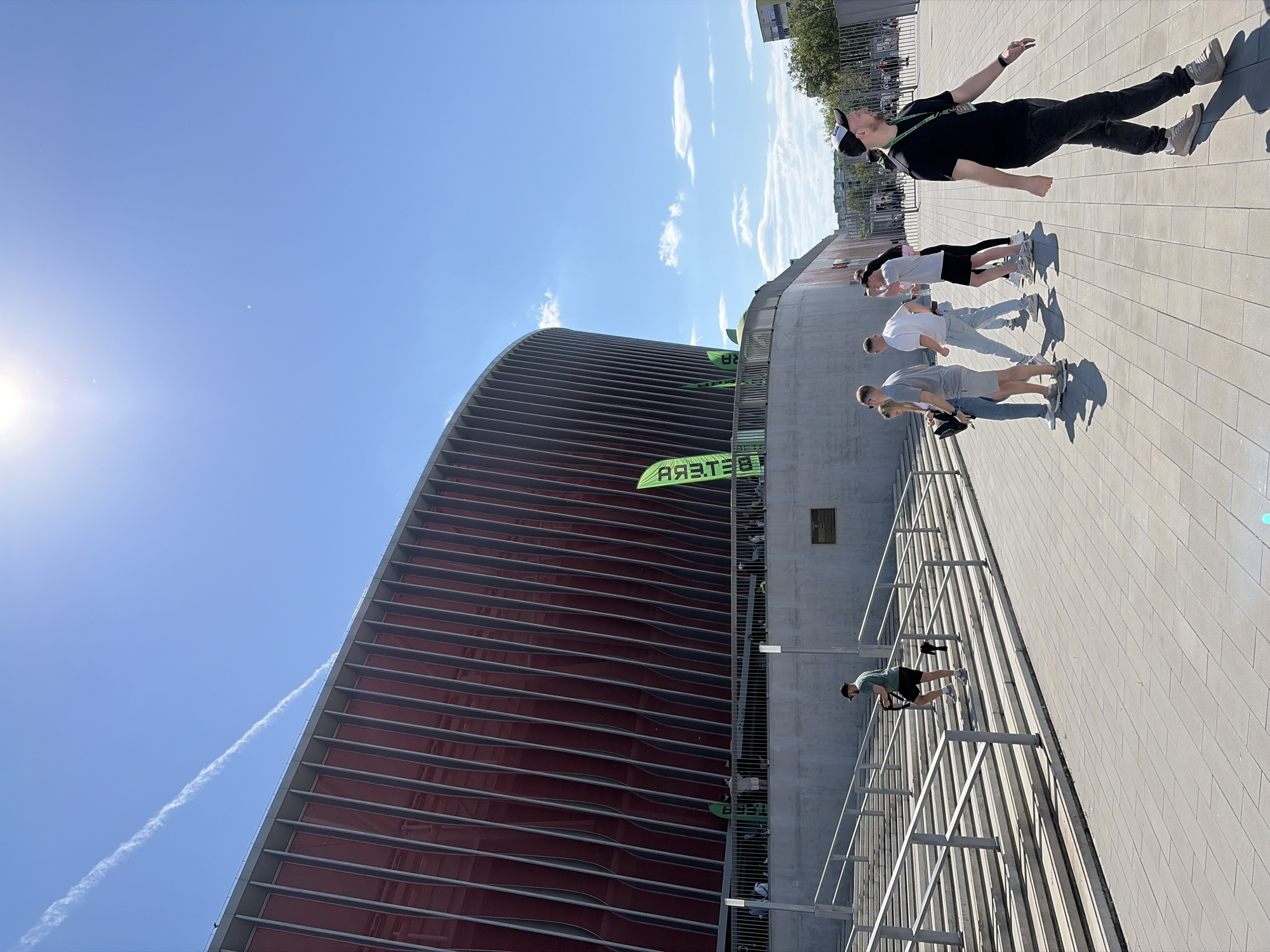 The image shows a group of people on a paved area in front of a large, curved building with a red and grey facade. The building has horizontal lines and is partially covered with green banners. The people are standing or walking. A metal railing and staircase are visible in the foreground. The sky is blue with some clouds. The photo was taken in Minsk, Belarus, near the Football Manege.