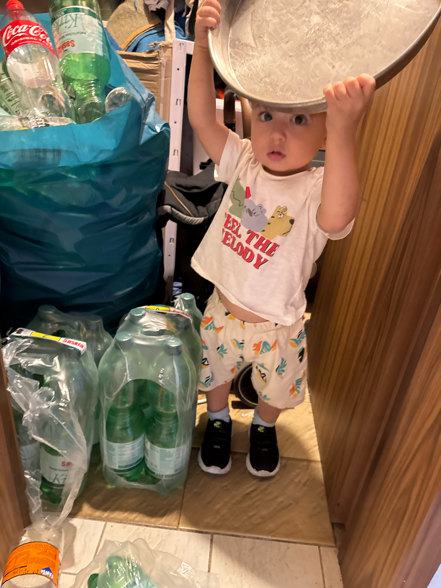 A young child is holding a metal tray over their head, standing in what appears to be a storage area. The child is wearing a white t-shirt with graphics of cartoon animals and the text "FEEL THE MELODY", and patterned shorts with a white background and colorful abstract designs. They are also wearing black sneakers with white soles and light blue socks.
To the child's left, there is a large, crumpled teal-colored bag filled with plastic bottles, including several clear Coca-Cola bottles and green bottles with labels. Next to the teal bag, there are several large plastic-wrapped multipacks of green bottles. Some of these bottles have yellow labels that read "Saskia" and "Ki". Further to the left, near the floor, is another plastic-wrapped bottle with an orange label.
The background consists of stacked items, including what looks like more bags, clothing, and possibly cardboard boxes. A white metal rack is visible behind the teal bag. To the right of the child, a wooden cabinet or wall with vertical lines is present. The floor is tiled in a beige color. The scene is indoors, suggesting a residential or storage space. No specific landmarks indicating the city of Überherrn, Germany, are visible in the image.