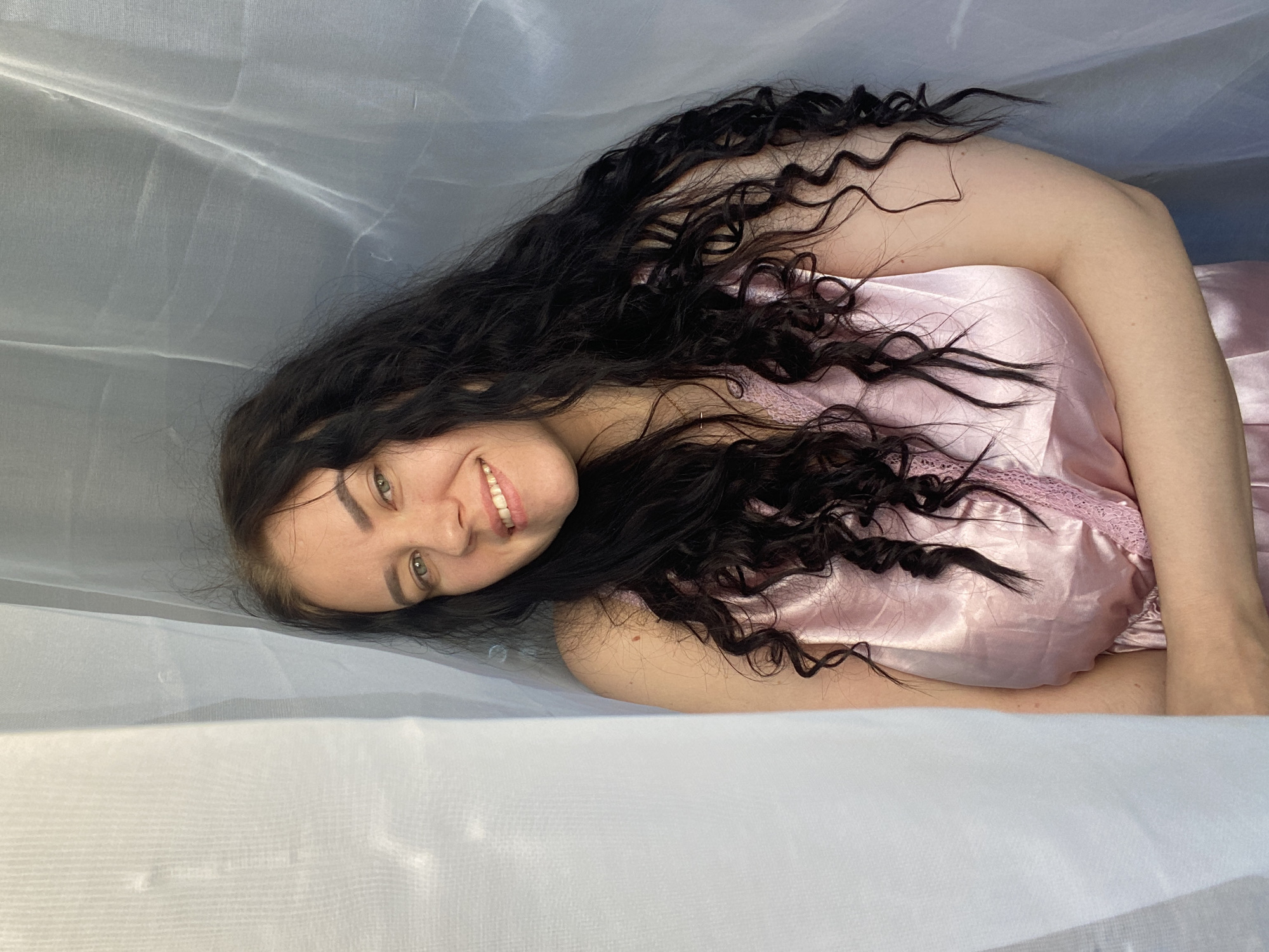 A woman lies sideways, head angled to the left, beneath a translucent white fabric. She has long, dark, curly hair and wears a pink satin garment with lace detail. Her eyes are green, and she is smiling, revealing her teeth. The location is not discernible from the image.