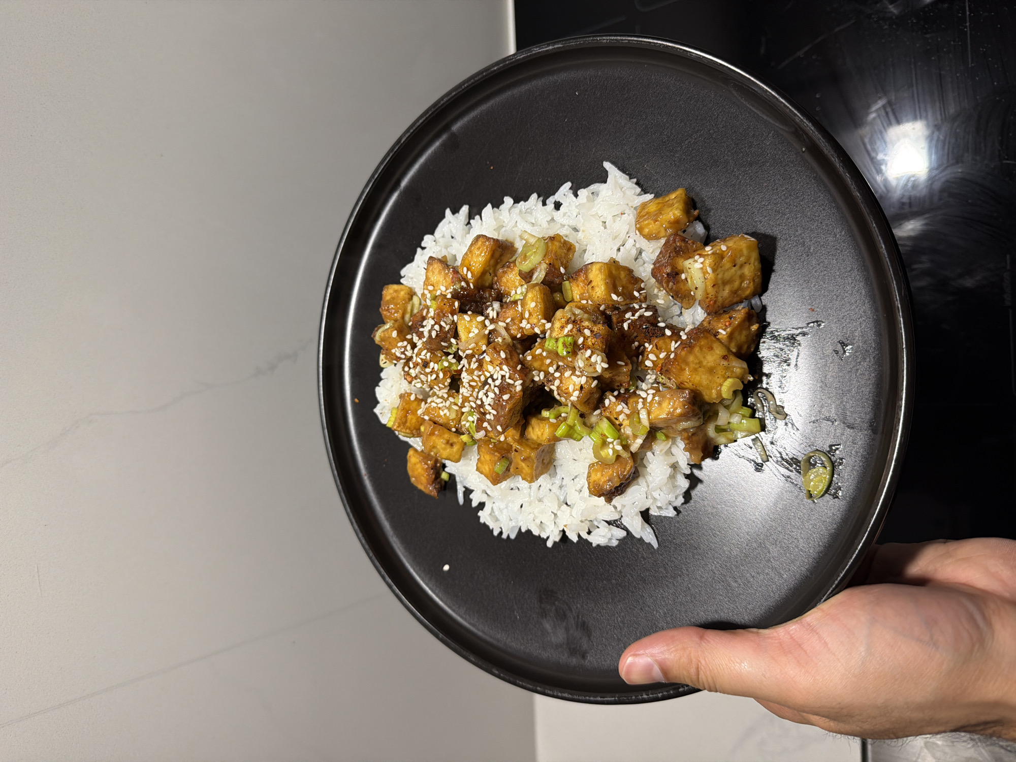 A person holds a black plate with a serving of white rice topped with small, cubed pieces of what appears to be glazed tofu. The tofu is sprinkled with sesame seeds and garnished with chopped green onions. The background includes a white countertop on the left and a black surface, possibly a stovetop, on the right. The location is not identifiable from the provided image.