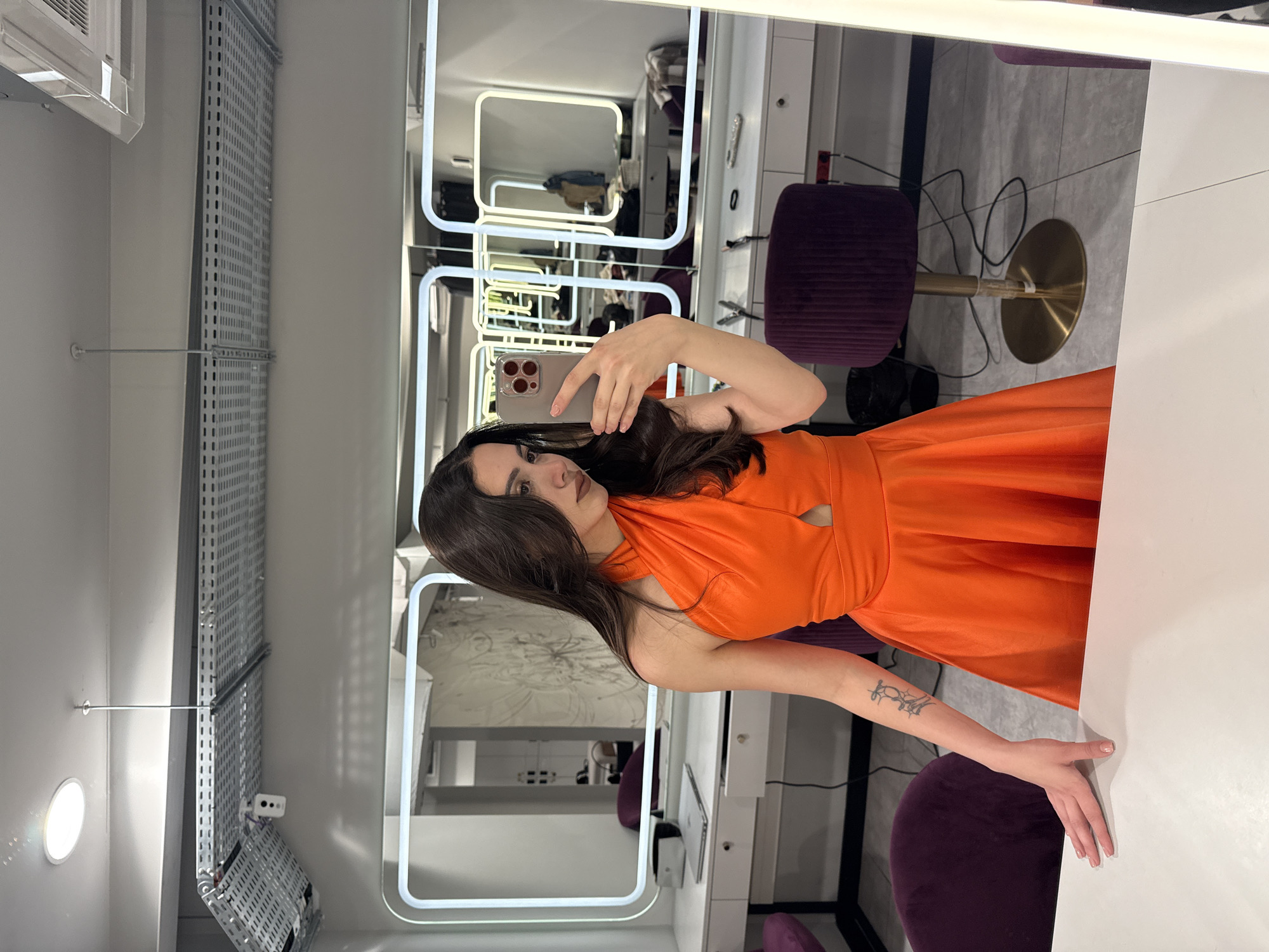 A young woman is captured taking a mirror selfie inside what appears to be a modern hair salon, beauty studio, or dressing room, located in Yerevan, Armenia. The original image is rotated 90 degrees clockwise.
The central subject is a woman with long, dark hair, wearing a striking orange halter-neck dress featuring a cutout detail at the chest. She is holding a smartphone, likely an iPhone, with a light-colored case, using it to photograph her reflection in one of the multiple illuminated mirrors. Her expression is neutral and composed as she poses for the selfie. A tattoo is visible on her right forearm.
The interior space has a clean and contemporary design, characterized by white walls and extensive white countertops or dressing tables. The flooring appears to be grey tile, visible in various reflections. Several large, rectangular mirrors dominate the scene, each framed with bright LED strip lighting, which suggests a professional setting for makeup or hairstyling.
Reflected in these mirrors are elements of the room's furnishings, including multiple purple velvet-upholstered stools or chairs with shiny brass bases. Additional visible details include an air conditioning unit mounted on a wall, a long perforated metal panel (possibly for shelving or utility organization), a small circular wall light, and various wires or cables on the floor. A small white dispenser or holder is attached to one of the tables. The overall ambiance is bright and well-lit by the internal lighting. No discernible text is visible in the image.