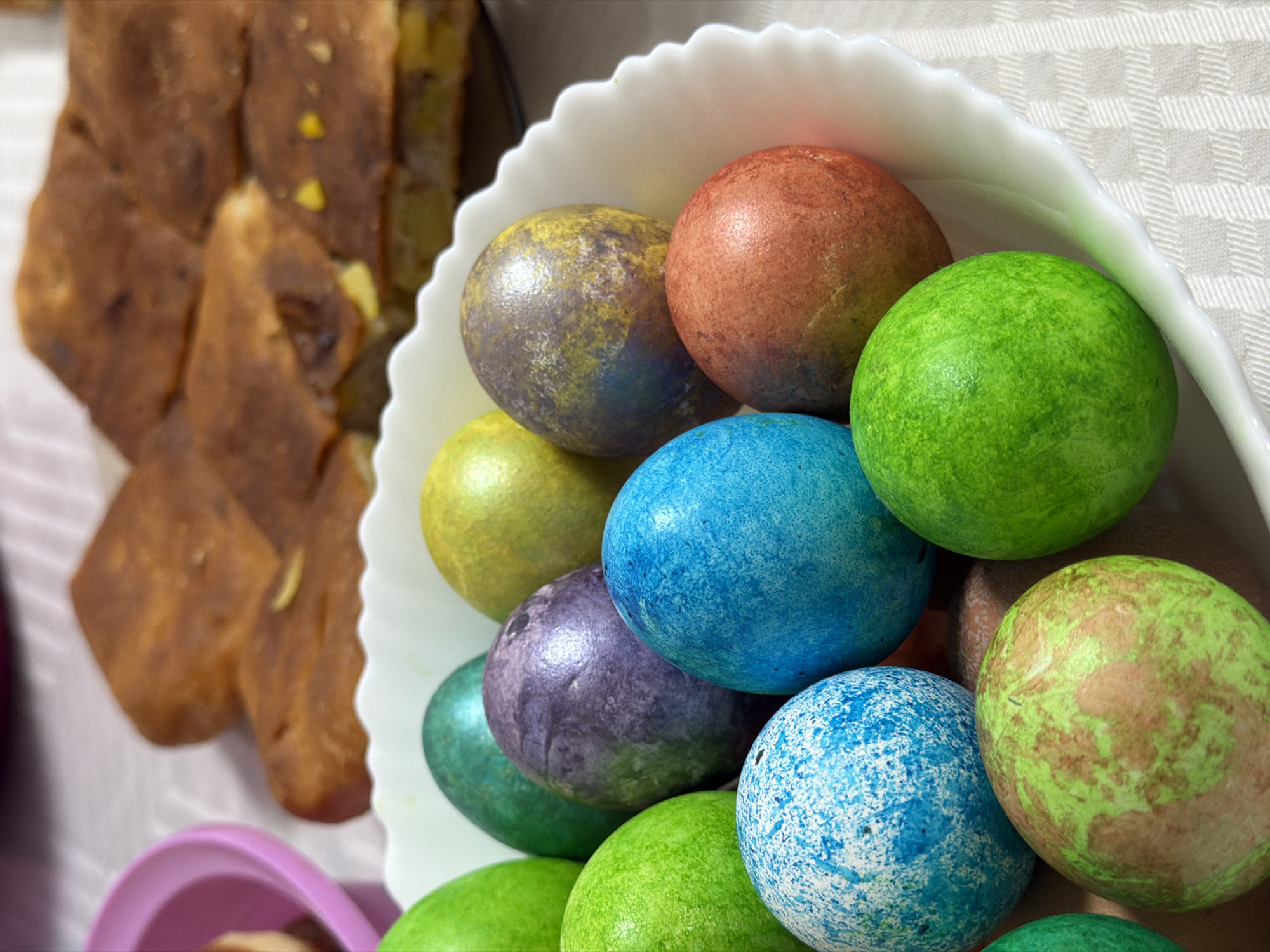 A close-up shot displays a white, scalloped bowl filled with colorful Easter eggs. The eggs are dyed in various shades, including blue, green, purple, orange, and yellow, with some having mottled or speckled patterns. To the left of the bowl, there is a stack of golden-brown pastries or breads. The background is a white and gray checkered surface. The image appears to be taken in Chaikovsky, Russia, but the precise interior location is not identifiable from the image alone.
