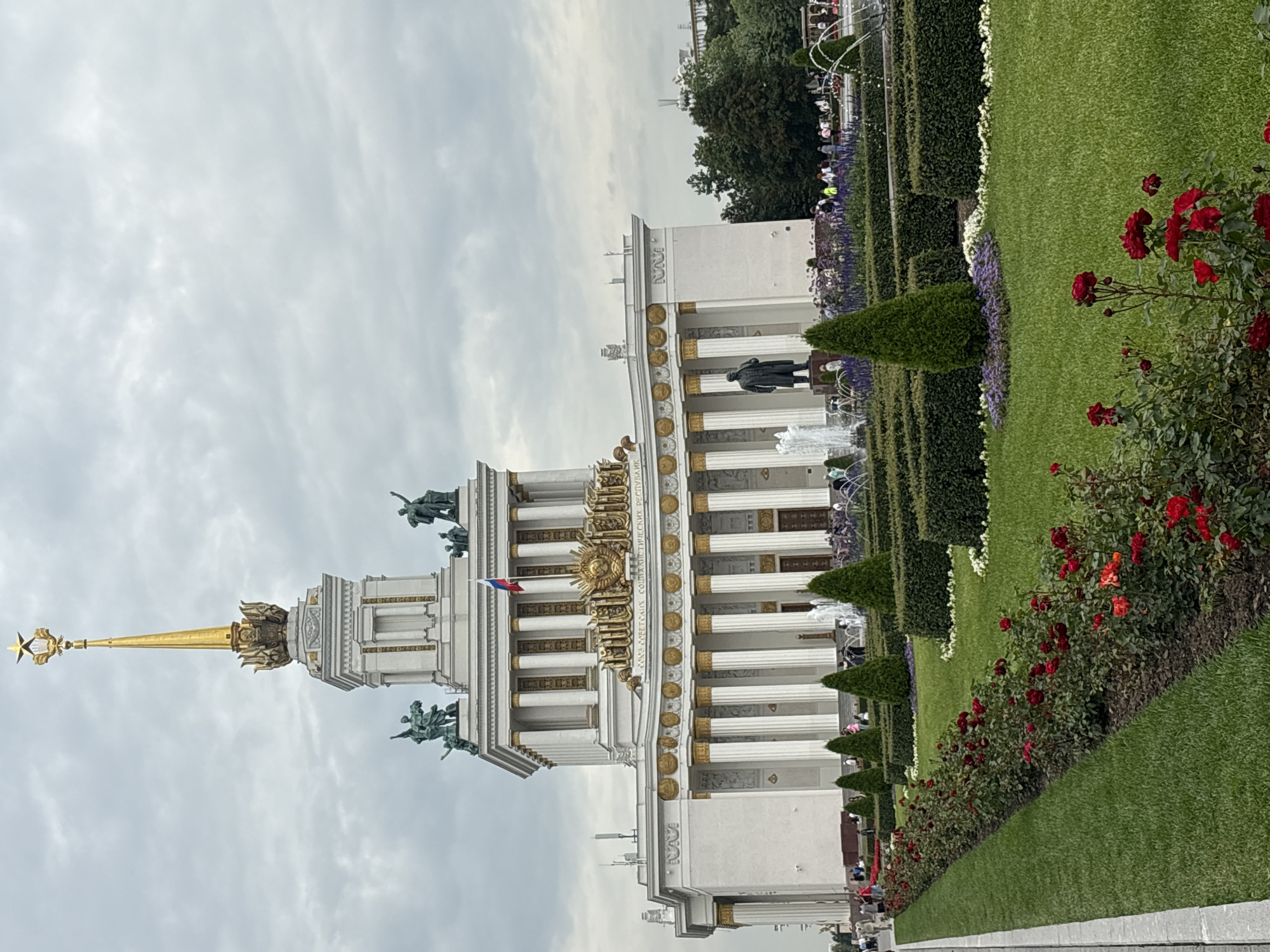 This is a photograph of the main pavilion at VDNKh (Exhibition of Achievements of National Economy) in Moscow, Russia. The pavilion is a grand white and gold building with a tall, ornate spire topped with a star. The facade features a colonnade of white columns with gold capitals, decorative friezes, and sculptures. In front of the pavilion, a statue of Lenin stands near several fountains and meticulously maintained gardens. The gardens are planted with rows of red roses, manicured hedges, and conical evergreen trees. People are visible walking in the background, and a Russian flag flies on a pole on the building. The sky is overcast with gray clouds.