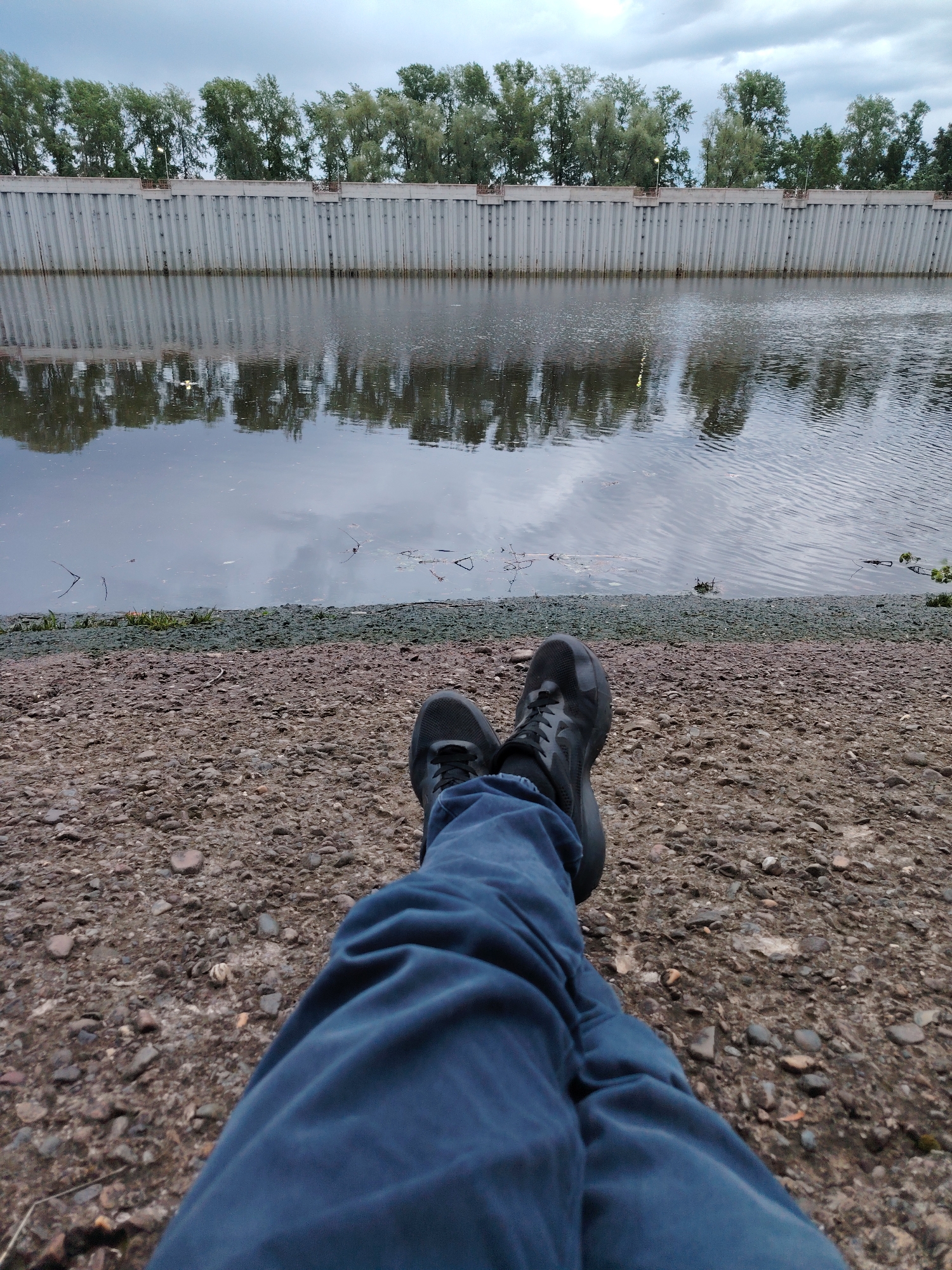 A first-person perspective photograph shows a person sitting on a rocky shoreline near a body of water. The person is wearing blue jeans and black sneakers. The background features a concrete retaining wall with trees lining the top and their reflections visible in the water. The location is likely in Chaikovsky, Russia, as it matches the typical landscape of the city. The scene appears calm and peaceful.