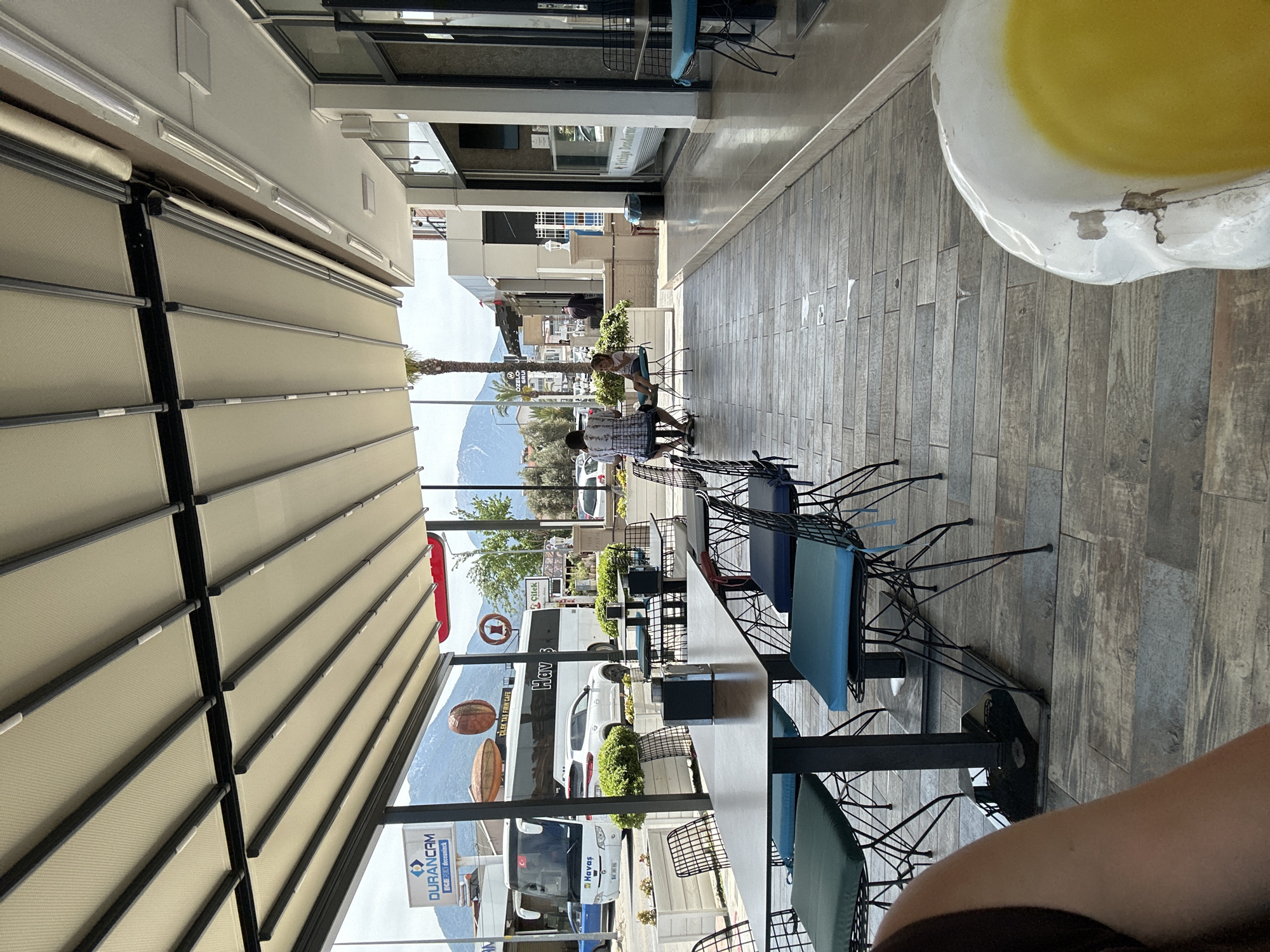 A restaurant in Fethiye, Turkey, showcases tables with blue chairs under an awning. Through the open-air structure, mountains are visible. People are sitting at tables further away. Signs and vehicles are visible through the opening.