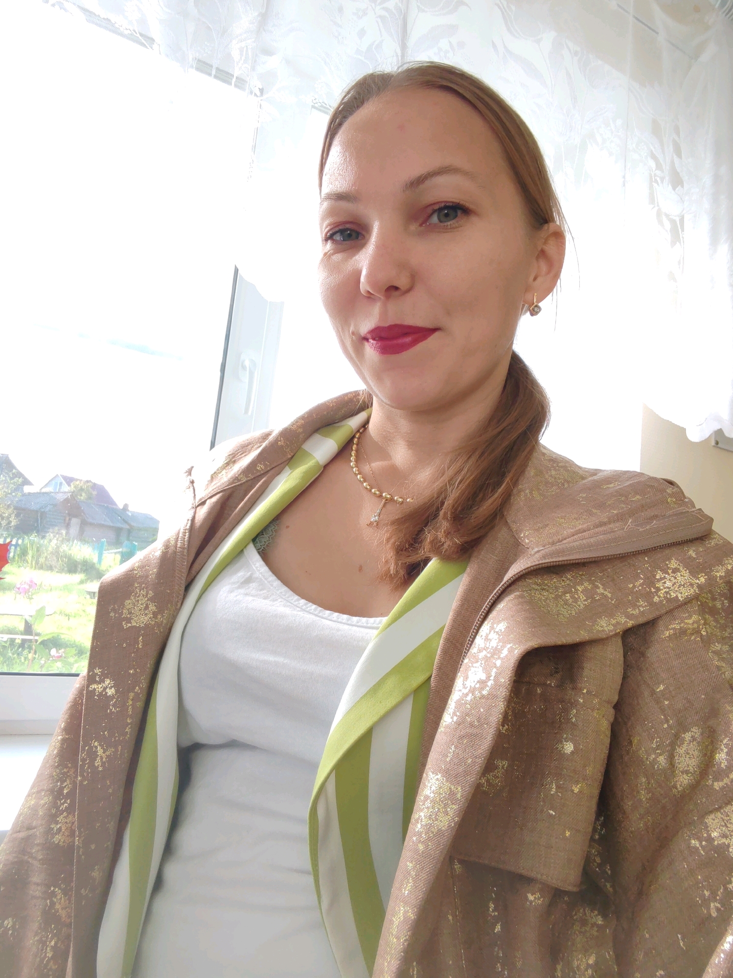 A woman with blonde hair pulled back in a ponytail and wearing a gold-flecked brown jacket over a white top smiles at the camera. She has on red lipstick and small gold earrings. The background is a window with white lace curtains, and through the window, a rural landscape with houses and trees is visible. The image appears to be taken indoors, near a window, in a rural setting.