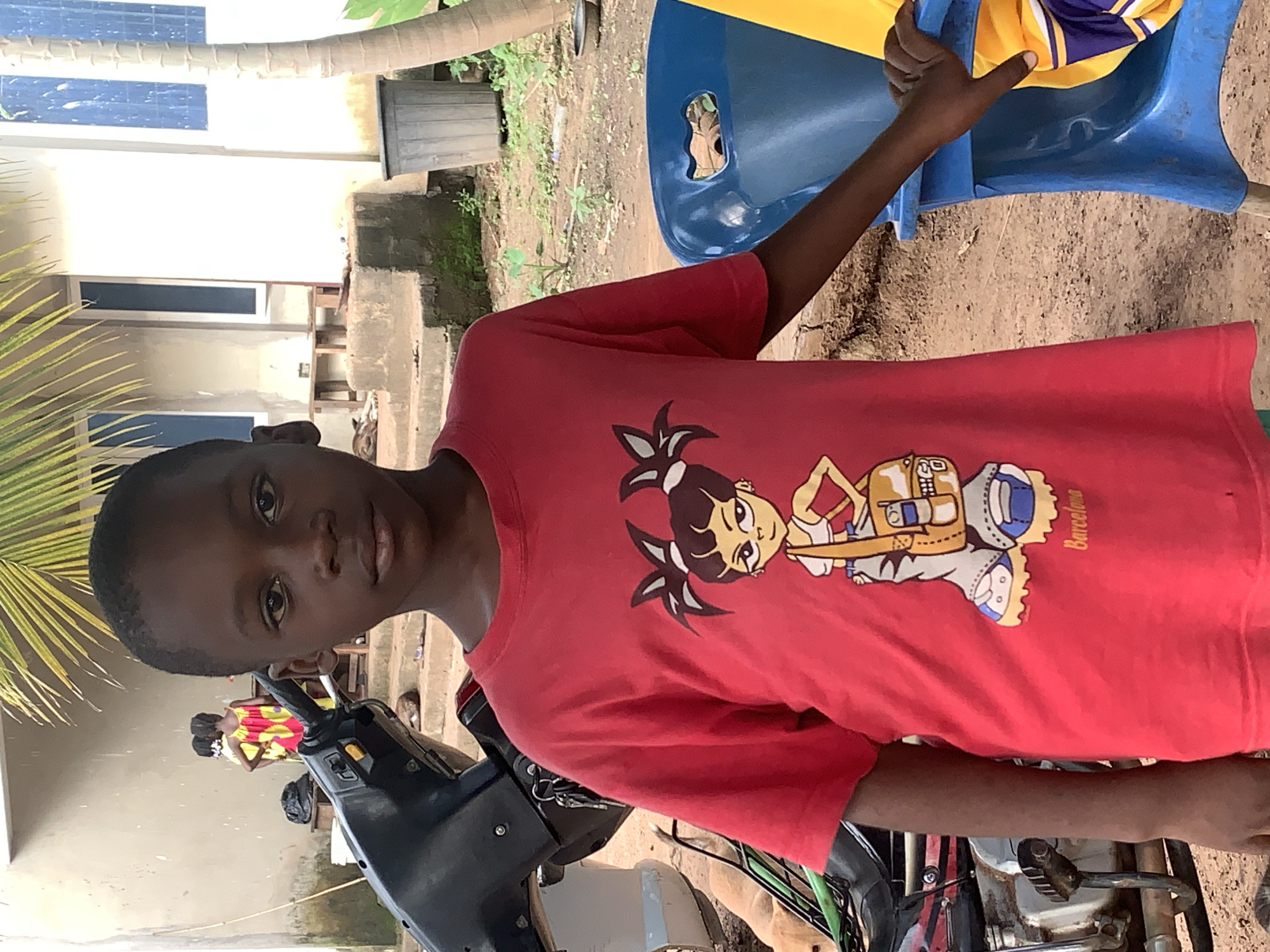 A young Black boy stands in a dusty outdoor area, positioned slightly right of center. He is wearing a bright red t-shirt with a graphic of a cartoon character in a sporty outfit. The boy's gaze is directed forward, and his expression is neutral. His left arm rests on a blue plastic chair, and his right arm is down by his side.
In the background, a light-colored building with windows is visible. To the left of the boy, a dark-colored motorcycle is parked. Further in the background, two young girls in colorful dresses can be seen standing near the building. The ground is covered in dirt and scattered debris. A large green palm frond extends from the upper left corner of the frame, casting a shadow. The overall environment appears to be a residential or communal outdoor space. The scene is located in Akwanga, Nigeria.