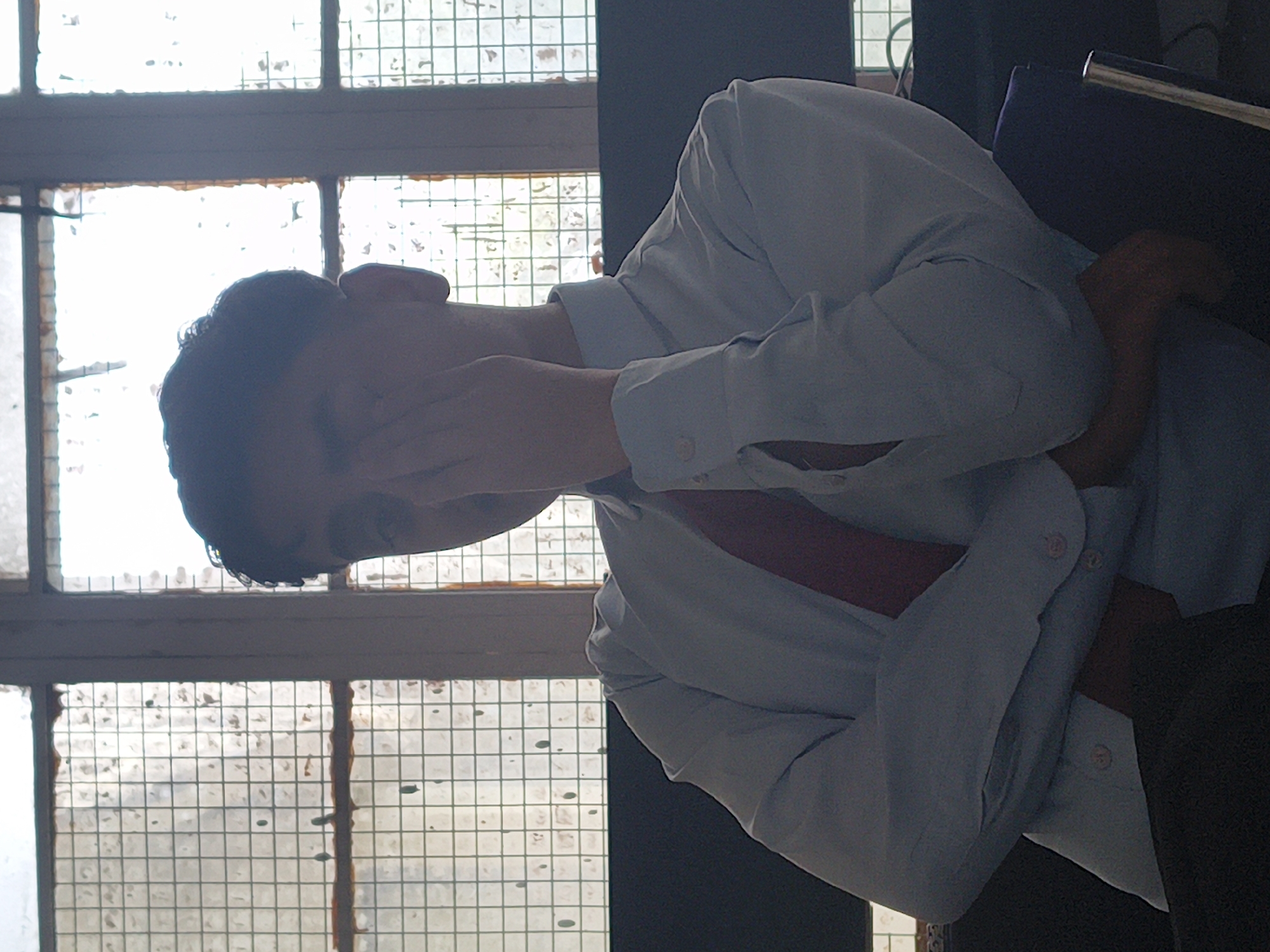 A young person, whose face is partially obscured by their hand, sits in front of a window with a grid pattern. The person is wearing a light blue dress shirt and a dark red tie. Their arms are crossed, and they are leaning back. The window behind them is backlit, obscuring the view outside and creating a silhouette effect. The scene is dimly lit. The specific location in Dharamshala, India, cannot be determined from the visual information provided.