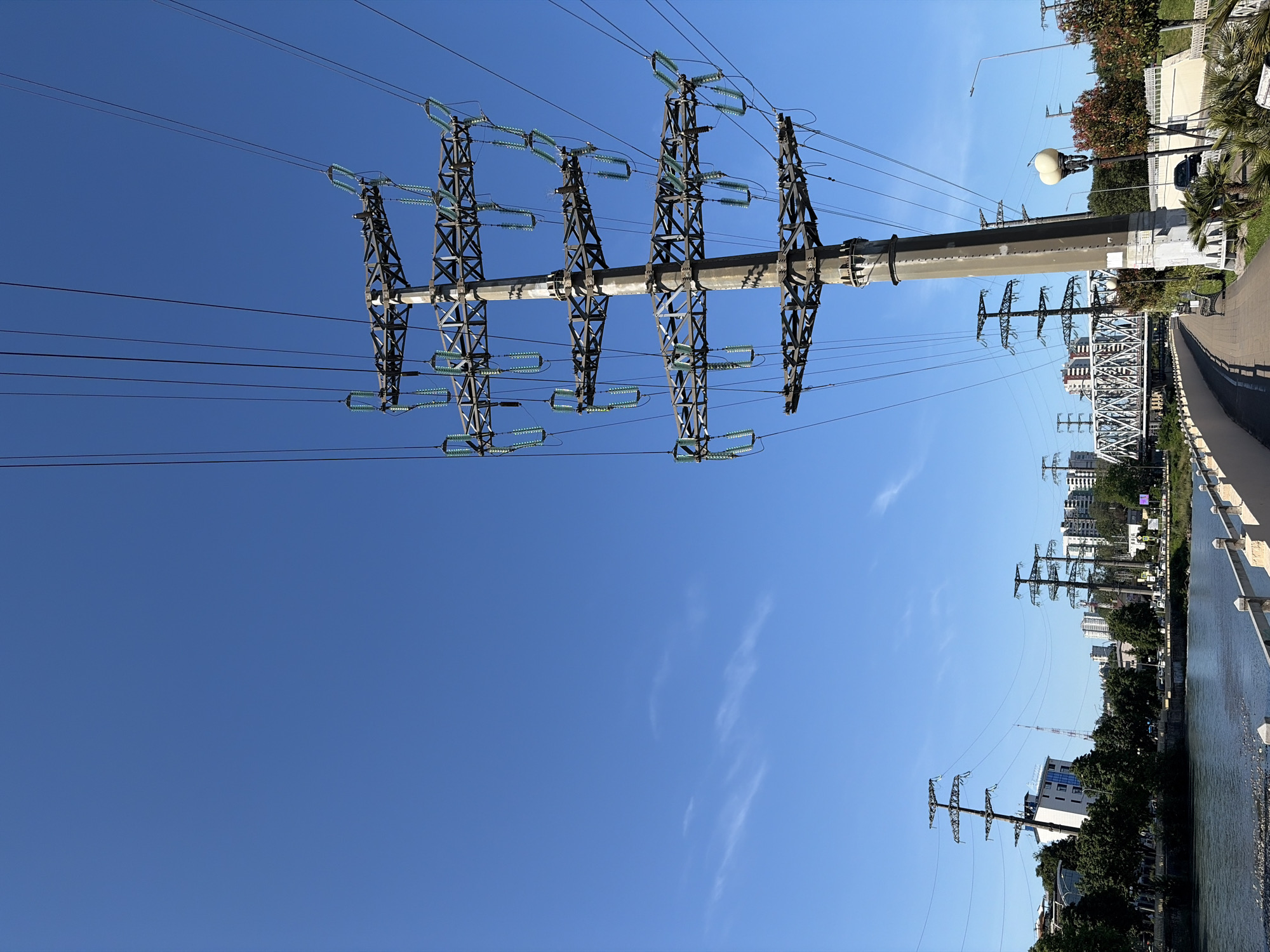 This media file captures a vibrant outdoor scene in Sochi, Russia, on a clear, sunny day. The prominent subject is a large power transmission tower (pylon) with a robust concrete base and intricate dark metal lattice structure, supporting numerous light-green glass or ceramic insulators from which multiple high-voltage power lines extend across the bright blue sky. A few wispy clouds are visible in the upper sky.
In the midground and background, to the right of the main tower, a paved promenade or walkway runs alongside a river, bordered by a white railing. Urban elements are scattered along this path, including decorative streetlights (one featuring a distinctive spherical light), green trees that appear to be a mix of deciduous and palm trees, and benches. A black car is partially visible behind some foliage, suggesting an active urban setting.
Further into the background, other smaller power pylons are visible, continuing the electrical infrastructure. A steel truss bridge spans the river, and beyond it, an urban skyline with various buildings completes the scene. The bright light and clear sky indicate it is daytime, likely mid-day. No people or specific activities are discernible, and no text is visible within the image. The scene depicts a blend of natural landscape with significant urban and industrial infrastructure along a riverfront.