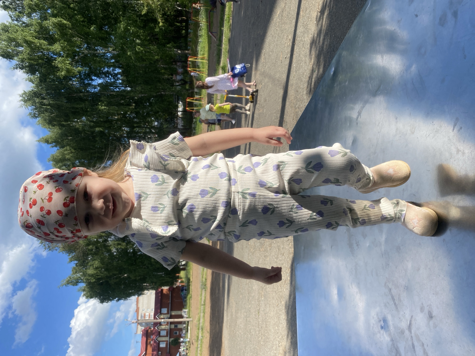 A young girl with blonde hair and wearing a light-colored jumpsuit with a cherry-patterned headscarf stands on a reflective surface. She is smiling and looking towards the camera. In the background, there are trees, a playground with other children, and buildings. The scene appears to be outdoors on a sunny day, possibly in a park or recreational area. The girl's playful expression and the presence of a playground suggest a lighthearted and leisurely activity. The reflective surface she's standing on might be part of a playground structure or a similar outdoor installation. The buildings in the background provide a hint of the urban or suburban setting.