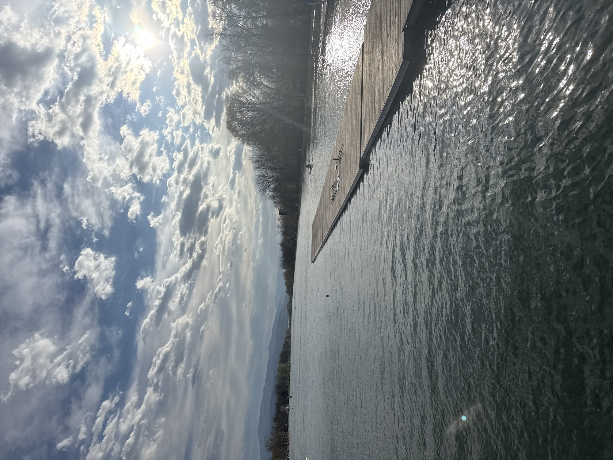 The image shows a body of water, likely a lake or river, with a wooden dock extending into it. The water surface is textured with ripples, reflecting sunlight. In the distance, a line of trees borders the water. The sky is partially cloudy with patches of blue visible.
The location could be Plovdiv Lake or the Maritsa River that runs through Plovdiv, Bulgaria. Further forensic details like GPS coordinates, objects with serial numbers, signs, or people with visible identifying markers would be needed to more precisely pinpoint the location and context.