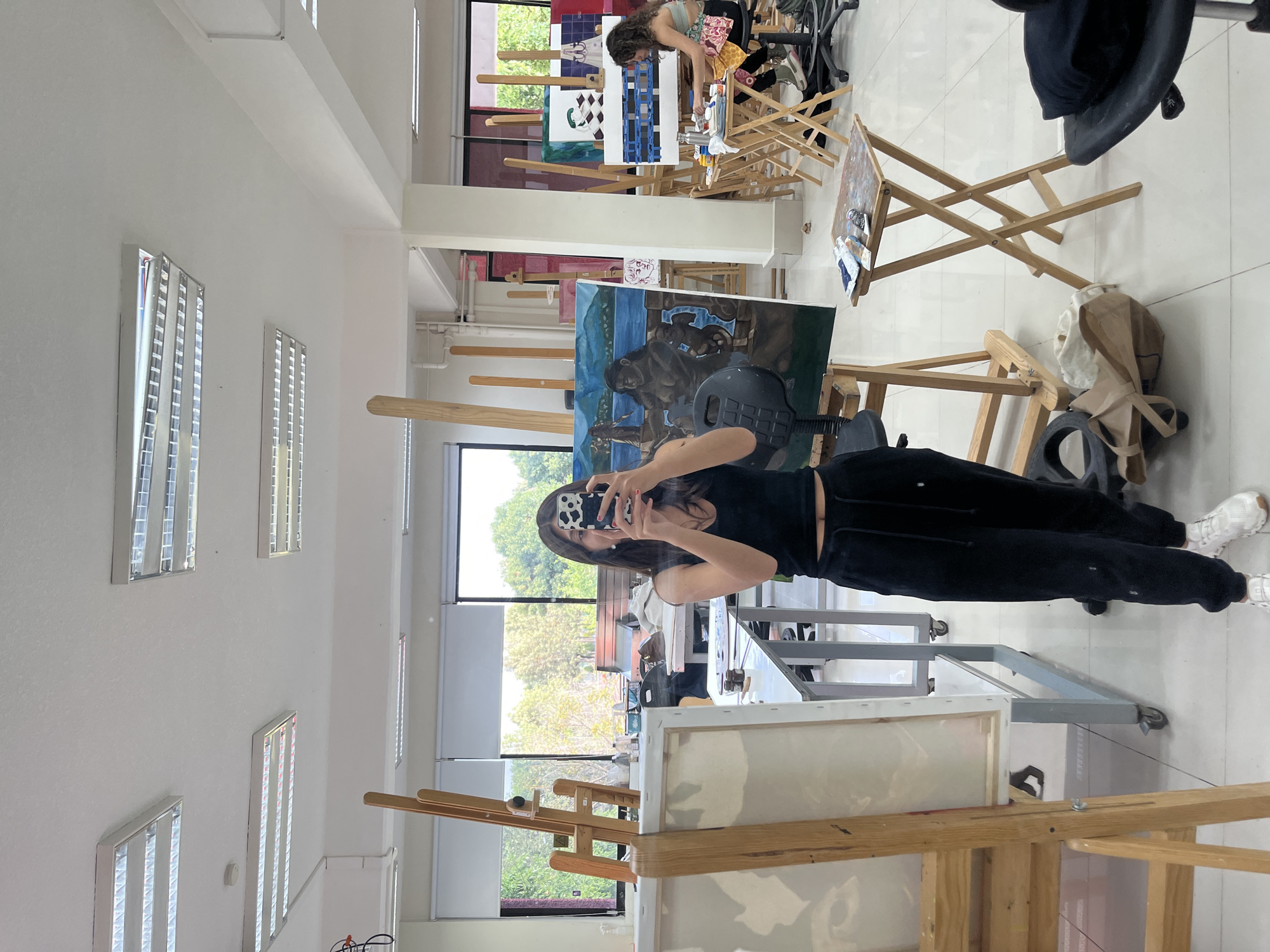 The image depicts an art studio setting, possibly within the city of San Andrés Cholula, Mexico. A woman is standing in the foreground, holding her smartphone and taking a photo. She wears black athletic clothes and white sneakers. Several art easels with paintings are visible, including one with a gorilla-like creature and blue landscape, and another one that is blank. Other students are present and working on art projects, visible in the background. The studio space has large windows providing natural light and views of the outside. The studio walls are white, with multiple fluorescent lights on the ceiling. Desks and other art supplies are scattered throughout the area. The floor is tiled in a light color.