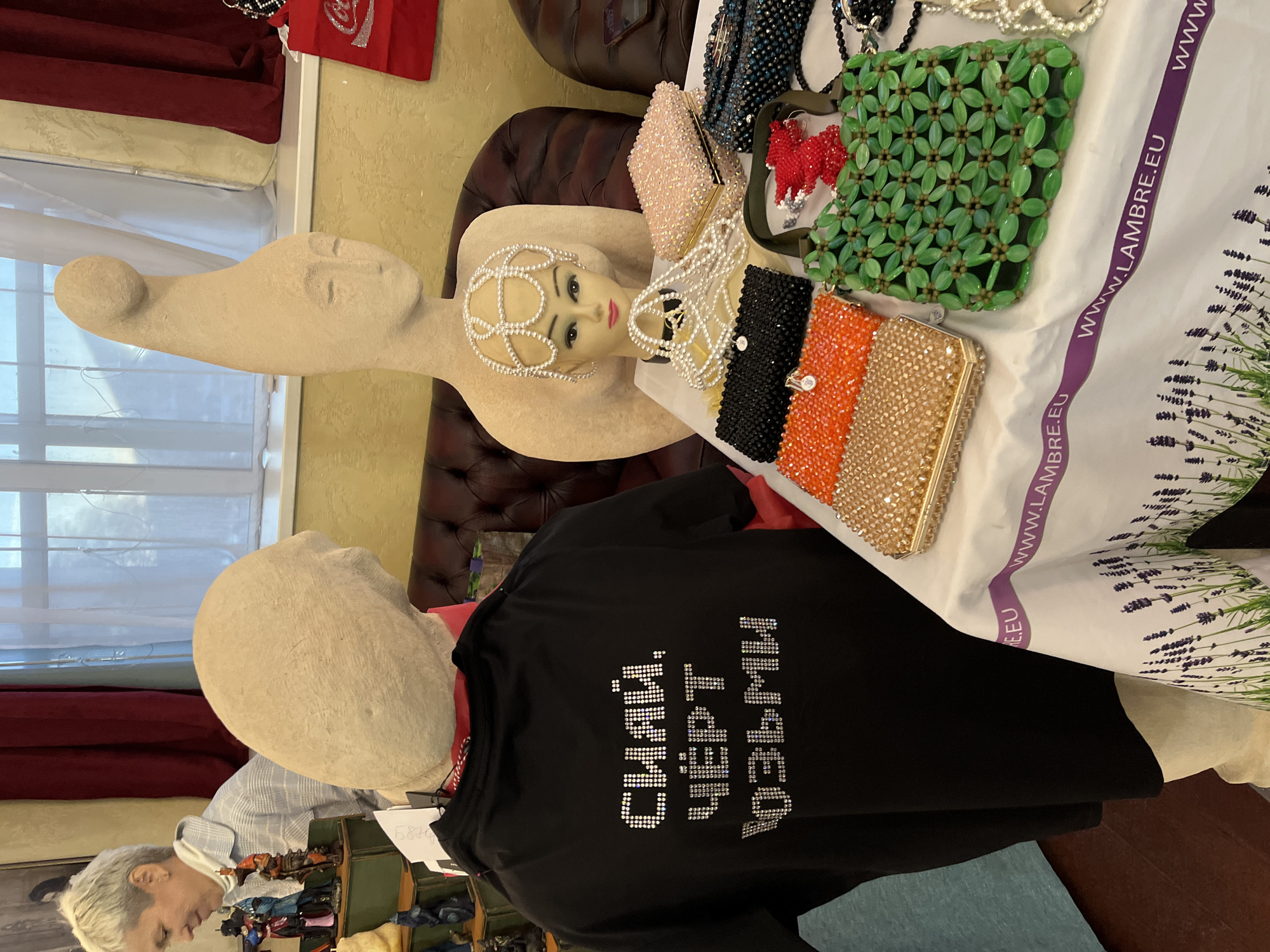 The image shows a display of fashion items. A mannequin head with a beaded headpiece rests against a tan sculpture. Several beaded clutch purses in various colors (pink, green, black/orange/gold, blue) are arranged nearby. A black t-shirt with Cyrillic text embellished with rhinestones is also on display. The text translates to "The smartest is always right." A white tablecloth with a lavender print and the website address "WWW.LAMBRE.EU" is visible. The background includes a leather chair, and a window. The location is likely a shop or market, but the exact location in Petropavlovsk-Kamchatsky, Russia, cannot be determined from this image alone.
