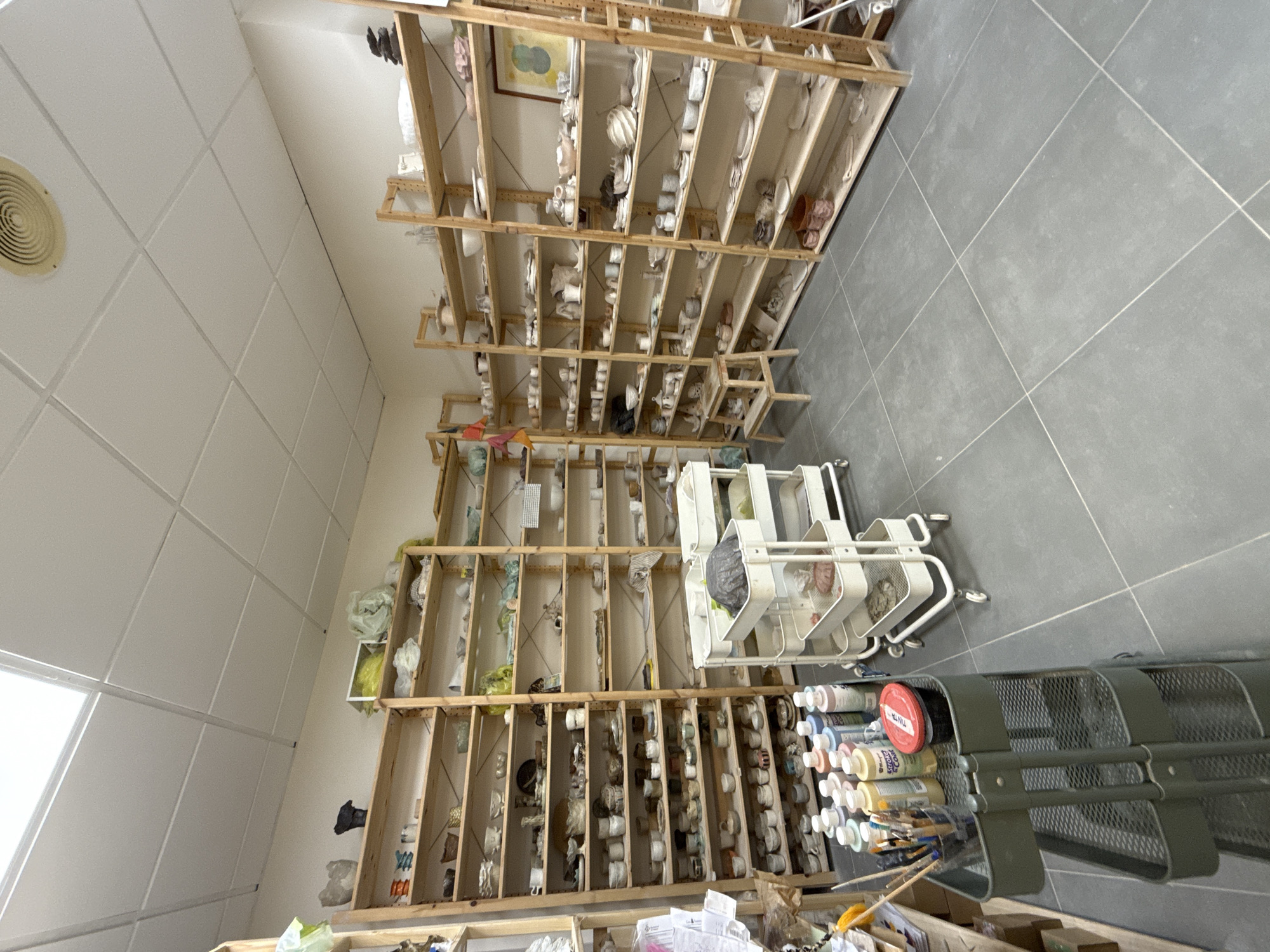 This is a medium-shot, eye-level, wide-angle photograph of a pottery studio, likely located in Mataró, Spain. The room is filled with numerous tall, wooden shelving units, packed with a variety of ceramic pieces in different stages of completion, as well as finished products. The shelves are organized and appear to contain bowls, cups, plates, and decorative items.
In the foreground, a grey, three-tiered rolling cart is visible. It holds a collection of art supplies, including bottles of colorful glazes, brushes in a container, and a red lidded jar. Another similar white rolling cart with three tiers is situated further back in the room, holding various plastic containers and what appear to be ceramic items or molds.
The walls are a neutral, light color, and the ceiling is a white drop ceiling with fluorescent lighting. A circular vent is visible on the ceiling in the upper right portion of the image. The floor is covered in large, grey square tiles with visible grout lines.
The overall impression is one of a functional and organized workspace dedicated to ceramic art. There are no people visible in the photograph.