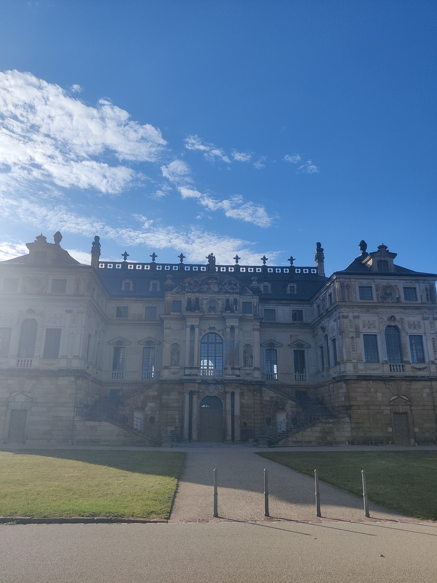 This is a full-frame, eye-level, outdoor, daytime photograph of the Baroque palace, Schloss Moritzburg, located in Saxony, Germany, near Dresden. The palace is situated on an island in the middle of a lake, and its distinctive architecture is the primary subject of the image. The palace features a grand facade with multiple stories, ornate detailing, and several prominent windows. A large, manicured lawn stretches out in front of the palace, with a pathway leading towards it. The sky above is a bright blue with scattered white clouds, and the sunlight casts shadows across the grounds. The scene conveys a sense of historical grandeur and serene natural beauty.