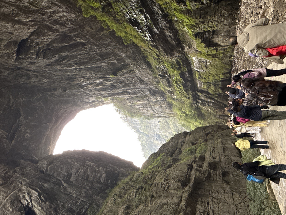 The image captures a detailed view of a natural geological formation within the Tiankeng-Difeng Scenic Area, Wulong District, Chongqing, China. The primary feature is a massive, naturally formed stone arch spanning the upper portion of the frame, through which a bright, distant vista of hazy green mountains and sky is visible. Towering, steep rock cliffs, characterized by dark gray, stratified layers and patches of green moss and vegetation, enclose the scene on both sides.
In the foreground and mid-ground, a group of approximately 15-20 individuals, appearing to be tourists, occupies a paved pathway constructed of irregular, light-colored flagstones. To the immediate right of the path, the ground transitions to an unpaved area composed of loose rocks and debris. Individuals are dressed in various casual outerwear, including jackets in shades of yellow, pink, blue, dark brown, and black. Multiple individuals in the center of the frame are holding mobile phones aloft, suggesting they are photographing or recording the natural structures. A man in the far left foreground wears a dark jacket and a blue backpack, walking away from the camera. Another man in a light brown hooded jacket is partially visible in the right foreground, also walking away, carrying a red garment over his left arm. The scene is illuminated by natural light, indicative of an outdoor environment.