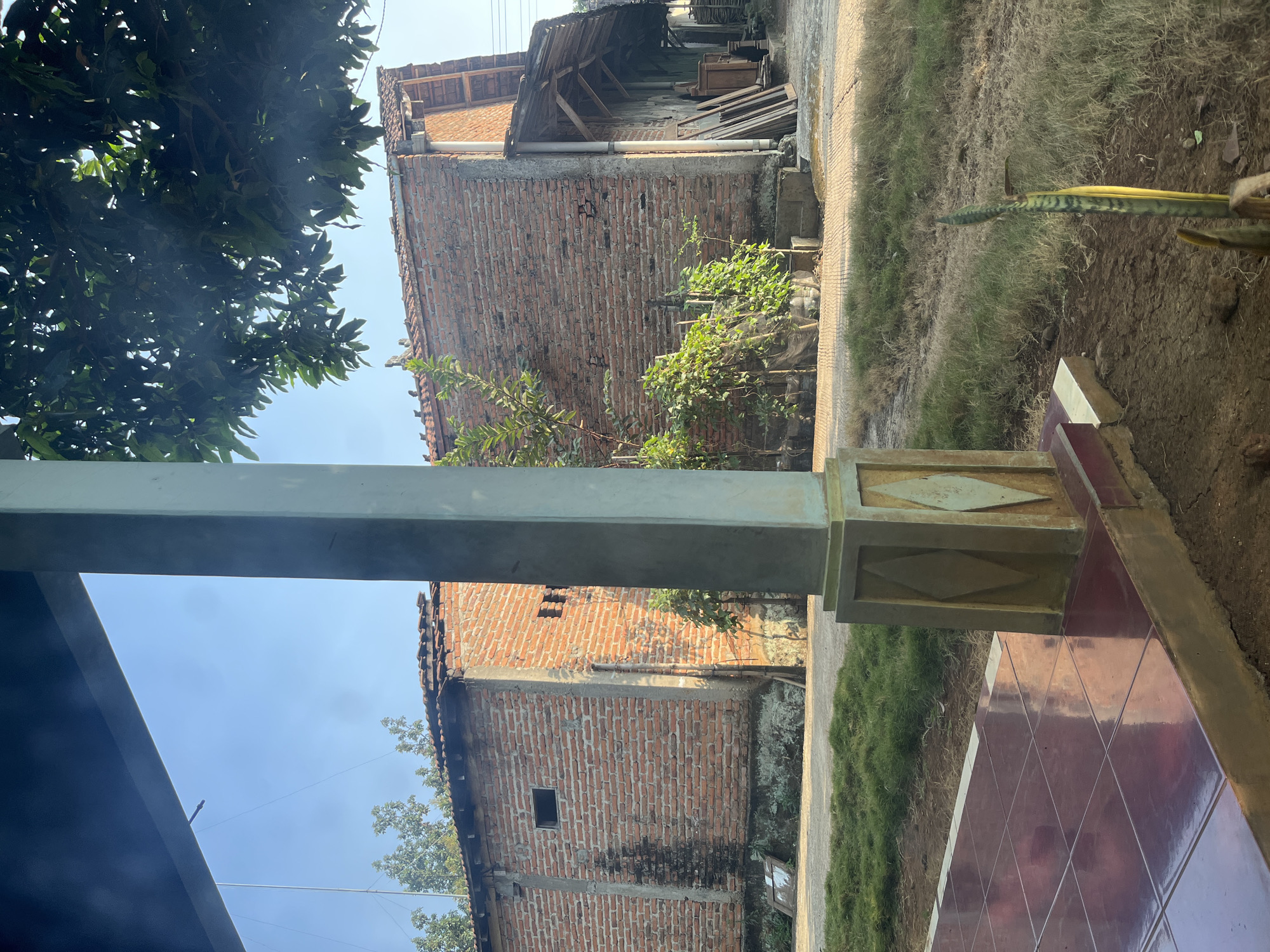 The image depicts an outdoor scene of a modest residential area in Glagah Kulon, Indonesia. In the foreground, a tiled patio with reddish-brown tiles leads to a textured pillar that is part of a covered structure. Beyond the patio, a patch of dry grass and dirt occupies the midground. Two simple brick buildings with tiled roofs are visible. The building on the left is a single-story structure with a small, dark rectangular window. The building on the right is also brick with a tiled roof and an overhang supported by wooden beams. Several green leafy plants are growing near the buildings. In the background, trees and the sky are visible. The overall impression is of a rural or semi-rural residential setting on a clear day, bathed in sunlight.