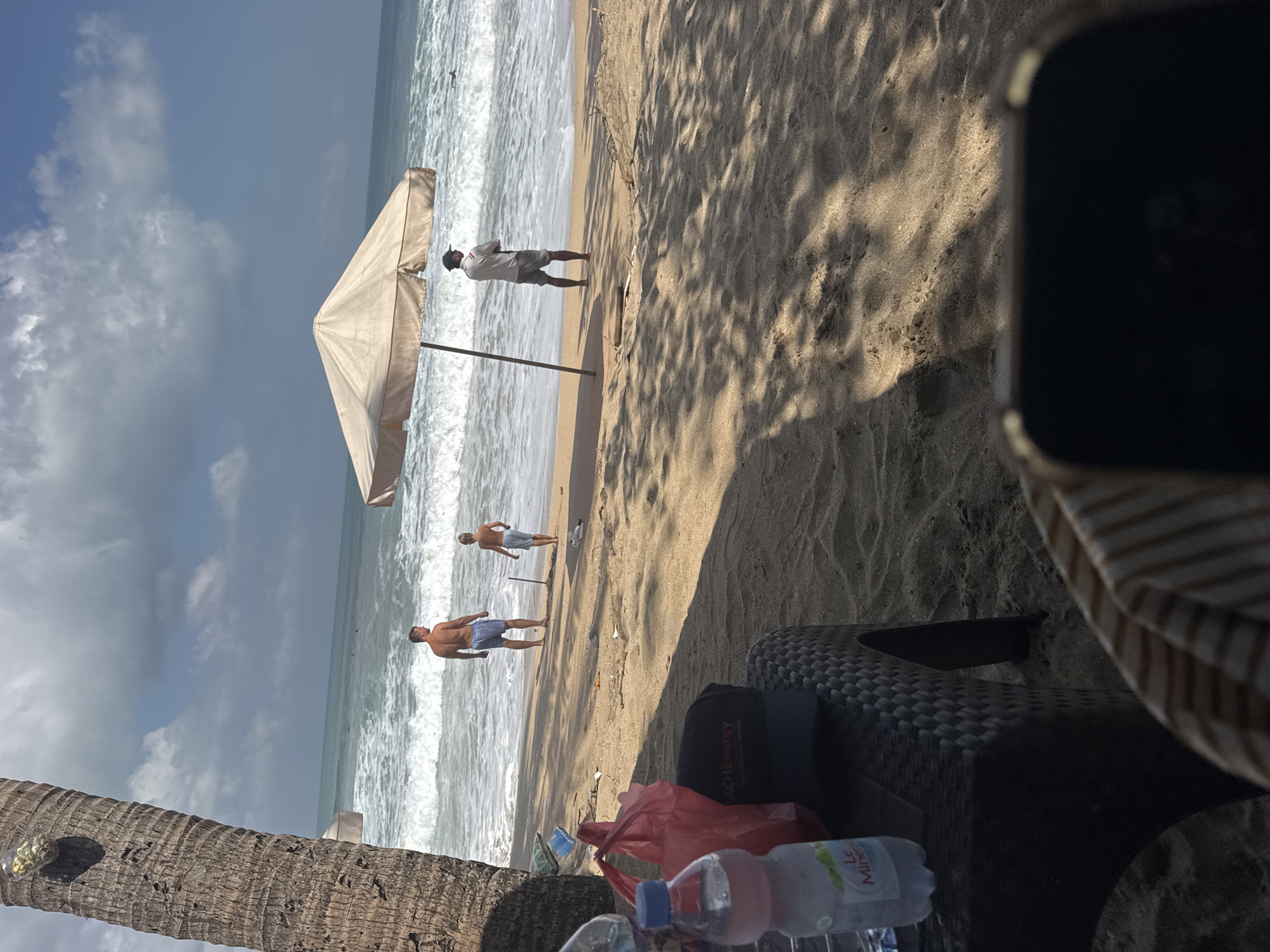 This is a medium shot of a tropical beach scene in Kuta, Indonesia. A tall palm tree trunk is on the left side of the frame, and its fronds cast shadows across the sandy beach. In the background, the ocean has moderate waves crashing onto the shore, with a few distant figures appearing to be in the water. A large, light-colored beach umbrella is set up on the sand. Two men and one boy are visible on the beach. One man, wearing blue shorts, stands near the water's edge looking out at the ocean, while a boy in light-colored shorts stands a short distance from him. Another man, wearing a white shirt and grey shorts, stands with his back to the camera under the umbrella, also looking towards the sea. In the foreground, a dark plastic chair is partially visible with a water bottle, a red plastic bag, and a dark baseball cap with the logo "ACH Energy" on it. The ground is sandy, with scattered debris and shadows from the palm tree and umbrella. The sky is partly cloudy with patches of blue.