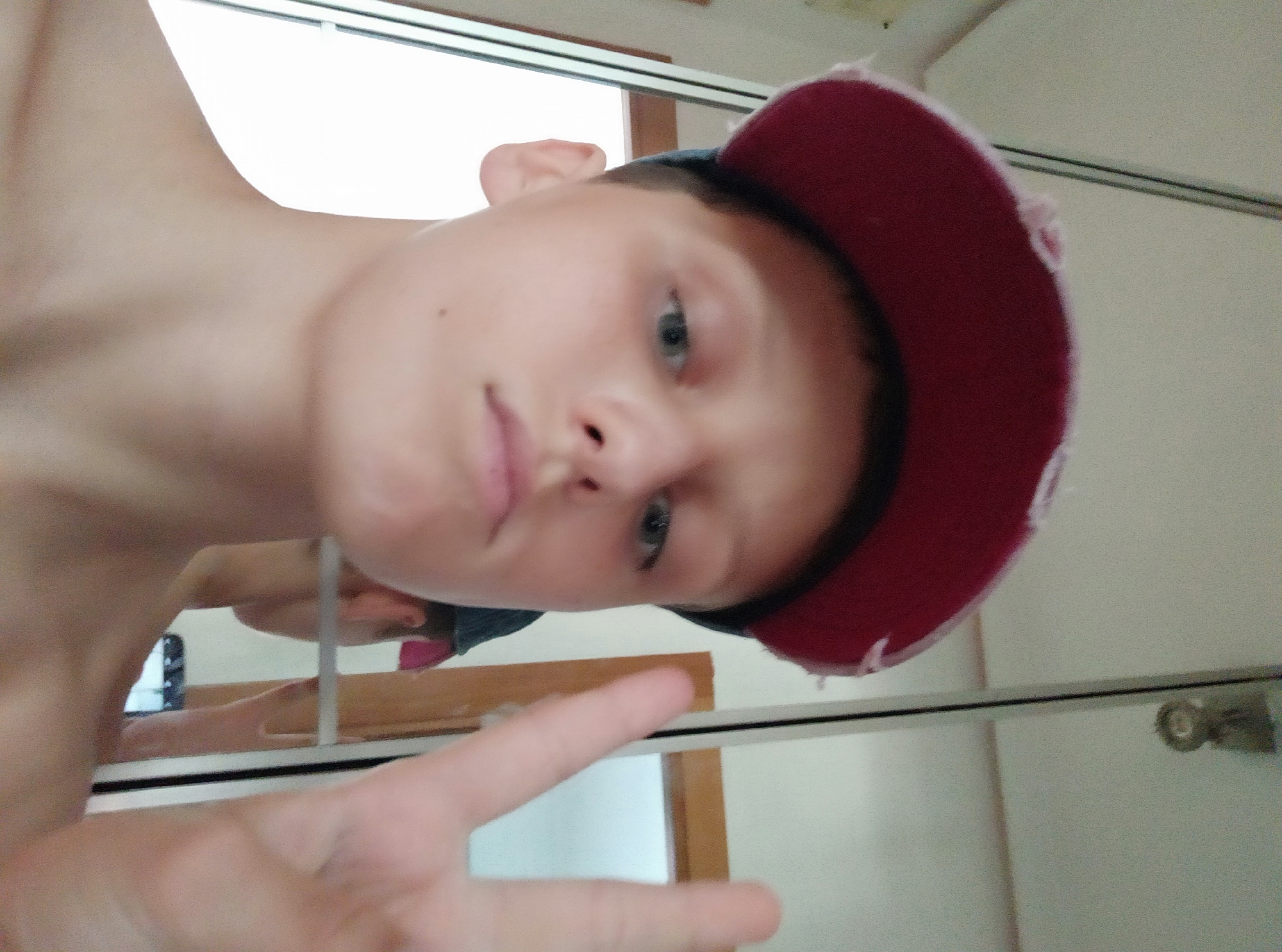 A young person, likely a boy, is captured in a close-up selfie. He is wearing a red baseball cap with a slightly distressed brim, tilted to the side. His face is the primary focus, with blue eyes looking towards the camera. His lips are slightly parted, and his skin appears fair. He is making a peace sign with his right hand, which is positioned in the foreground, slightly out of focus. In the background, a mirror reflects the scene, showing a glimpse of another person, possibly the same individual, and a door or frame. The environment appears to be indoors, with a plain wall and a light fixture visible in the reflection. The lighting is bright, suggesting natural light from a window or a well-lit room. The composition is informal, typical of a casual selfie.