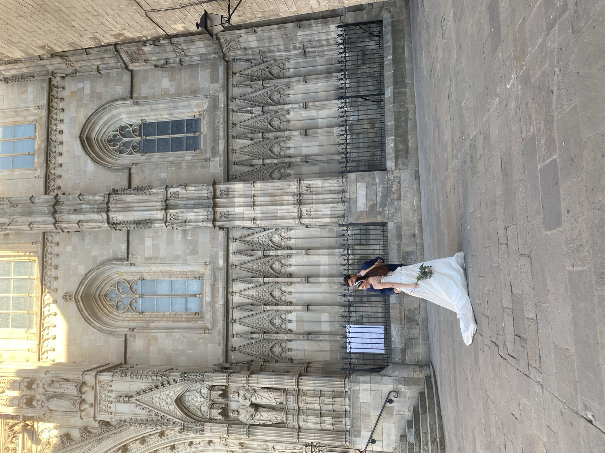 In Barcelona, Spain, a bride and groom stand on a stone-paved plaza in front of the Barcelona Cathedral. The bride, wearing a white wedding dress with a long train and a floral bouquet, stands with her back mostly to the camera, looking over her left shoulder. The groom, in a dark suit, stands behind her and to her right. Behind the couple is the imposing facade of the Barcelona Cathedral, featuring gothic architectural elements, ornate stone carvings, and arched windows. A black metal fence separates the plaza from the cathedral's base, and a set of stone stairs with a railing is visible to the left. Sunlight casts a warm glow on the upper portion of the cathedral building.