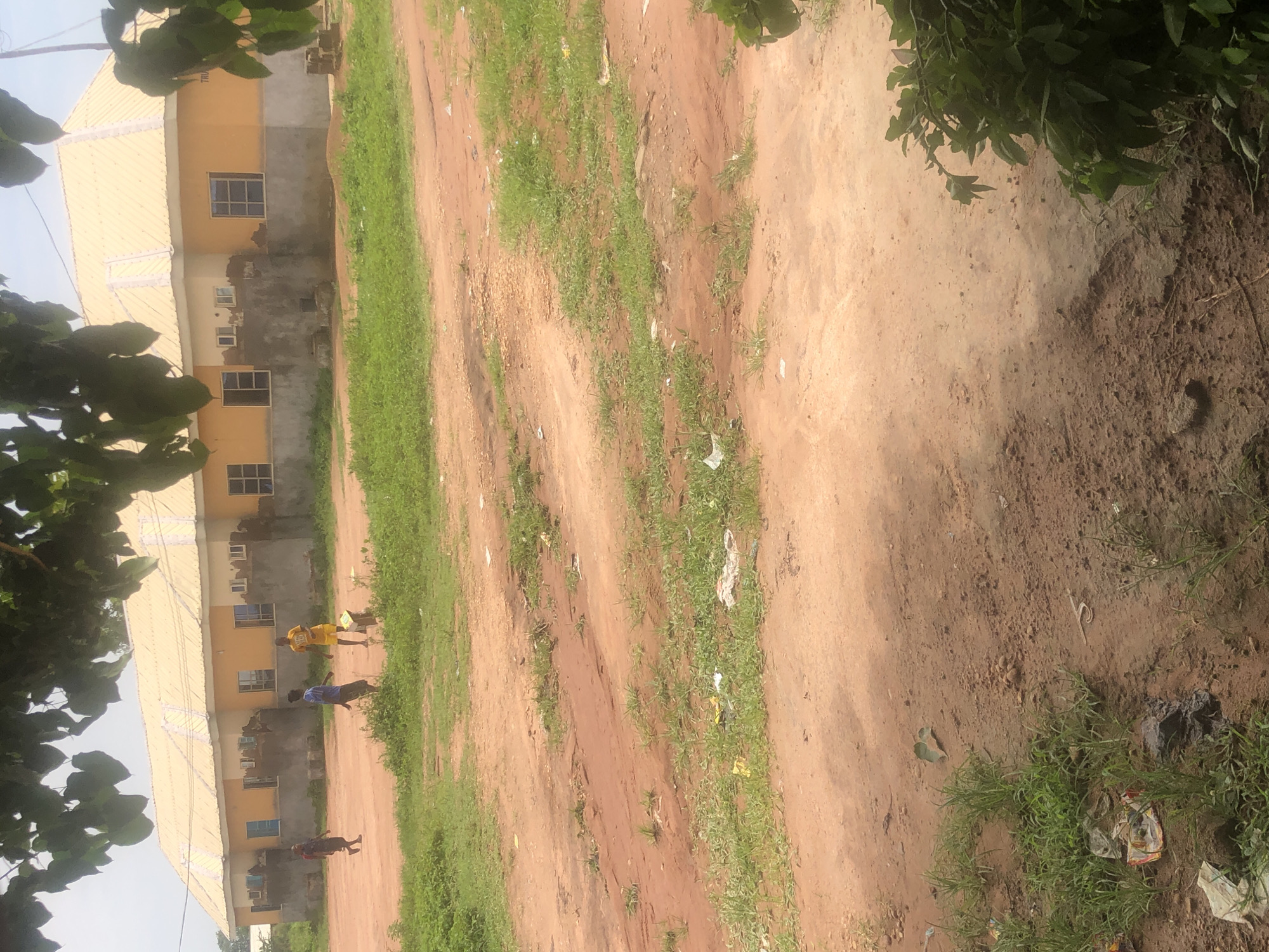 The image depicts an outdoor scene with a building and several individuals in what appears to be a construction or undeveloped area. The building is a single-story structure with an orange facade and a corrugated yellow roof. It has several windows, some of which appear to be boarded up or under construction. The ground is predominantly red dirt with patches of green grass and some scattered debris.
In the foreground, there is a dirt path or clearing with sparse vegetation. In the mid-ground, closer to the building, three individuals are visible. One person, wearing yellow shorts and a t-shirt, is standing near a small, light-colored object. Another person, wearing a blue shirt and dark shorts, is walking. The third individual, wearing a dark shirt and shorts, is also walking. The scene suggests a casual or possibly work-related activity in this location.
The environment appears to be an open space with hints of ongoing construction or development. The overall impression is one of a rural or semi-urban setting, characteristic of parts of Wukari, Nigeria. The lighting suggests it is daytime, possibly late morning or afternoon, with bright, overcast conditions.
