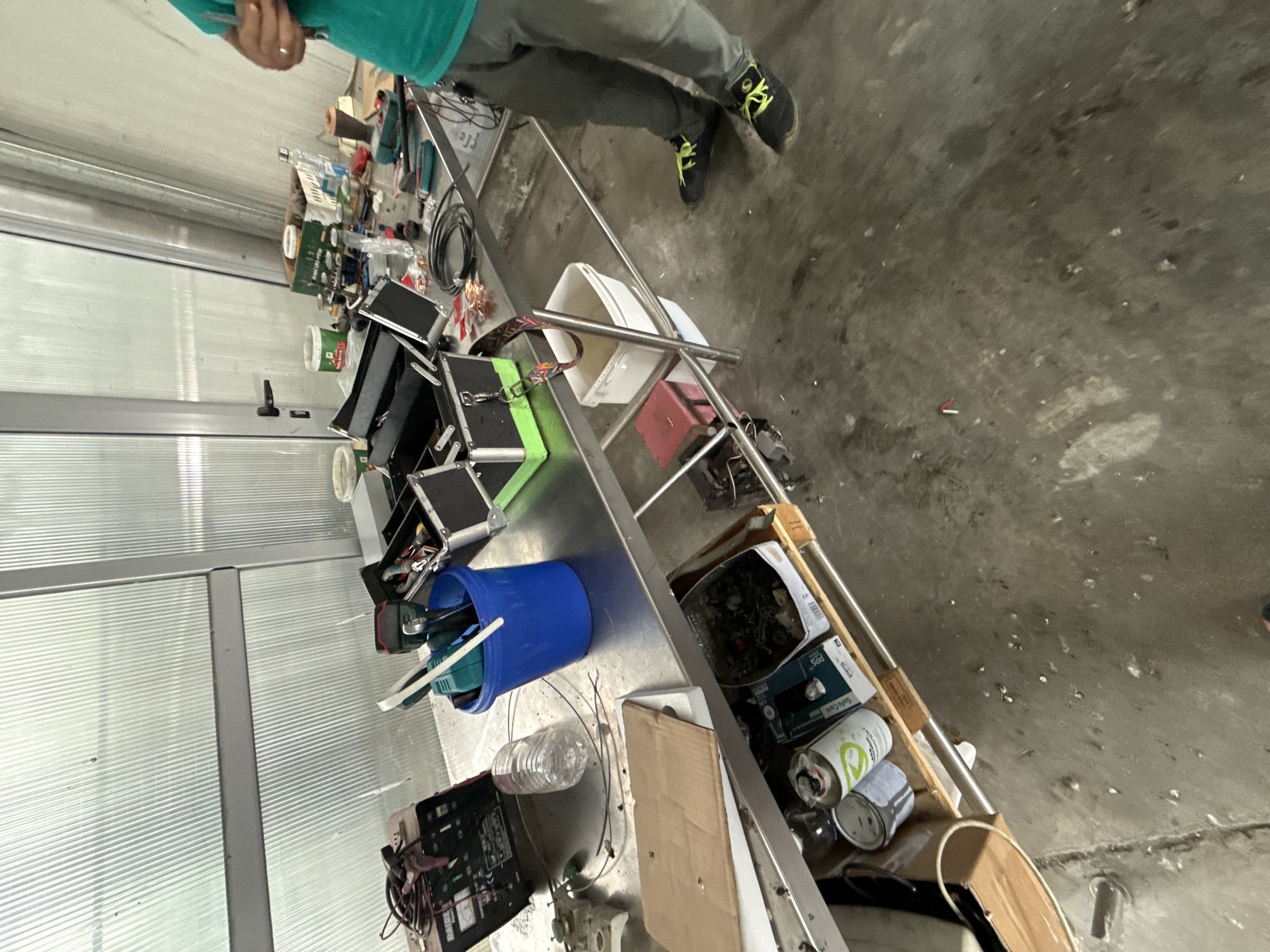 This media file depicts an indoor industrial or workshop environment, possibly located in Stara Zagora, Bulgaria. The central focus is a long, metallic workbench heavily laden with various tools, electronic components, and miscellaneous containers.
A person is partially visible in the upper left quadrant of the image, wearing a green t-shirt, grey trousers, and black sneakers with distinctive neon green laces. Their hands are seen near the top of the frame, suggesting engagement with the items on the table, though no specific activity is clearly discernible.
The workbench is crowded with a diverse array of objects: power tools including a green and a blue drill-like device, numerous circuit boards, a coiled black cable, and several metal cases that appear to be toolboxes or equipment storage. A blue plastic bucket is filled with tools, and a clear plastic water bottle rests nearby. A battery charger bearing the brand name "RAIDER" is also visible. Among other items are various cans, one of which is white and blue with "PPS" and "Safe Care" text, and another featuring a green logo. Additional containers, such as a white plastic tray, a red box, and cardboard boxes brimming with small parts, contribute to the cluttered yet functional appearance of the workspace.
The floor is a rough concrete surface, showing signs of wear and dirt, with some scattered debris. In the background, translucent, ribbed panels, likely forming walls or partitions, allow ambient light to illuminate the space, suggesting it is daytime. These panels imply a setting such as a warehouse, garage, or a temporary workshop structure. The overall impression is that of an active workspace used for repairs, assembly, or dismantling tasks.