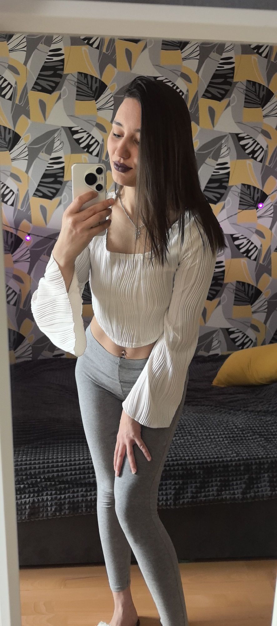 A woman is taking a selfie in front of a mirror. She is wearing a white, textured, cropped long-sleeve top with wide, flared sleeves, and grey leggings. Her hair is dark and straight, and she has dark lipstick on. She is holding a smartphone with a triple-lens camera. Behind her is a bed with a grey textured comforter and a yellow pillow. The wall behind the bed is covered with a patterned wallpaper in shades of yellow, grey, and black. There are also string lights with purple bulbs hanging on the wall. The floor appears to be wooden. The location is Unknown, Unknown.