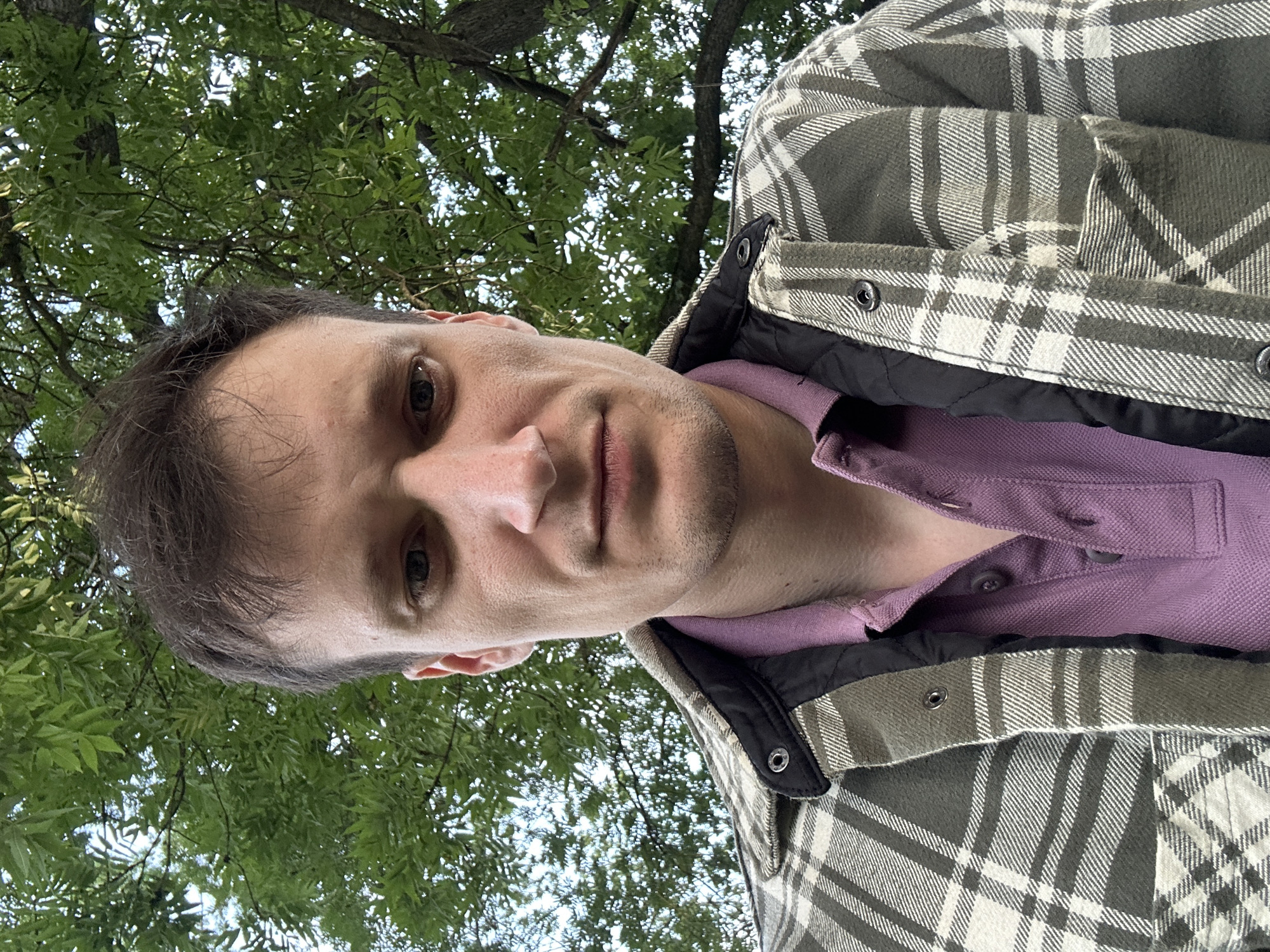 A medium shot selfie shows a man looking directly at the camera from a slightly upward angle. He has short, dark brown hair and a fair complexion. He is wearing a purple collared shirt under a plaid jacket with gray and white patterns. The background is dominated by lush green foliage and a tree trunk, suggesting an outdoor setting. The lighting is natural and appears to be daytime. No specific location in Makiv, Ukraine is discernible from the image.
