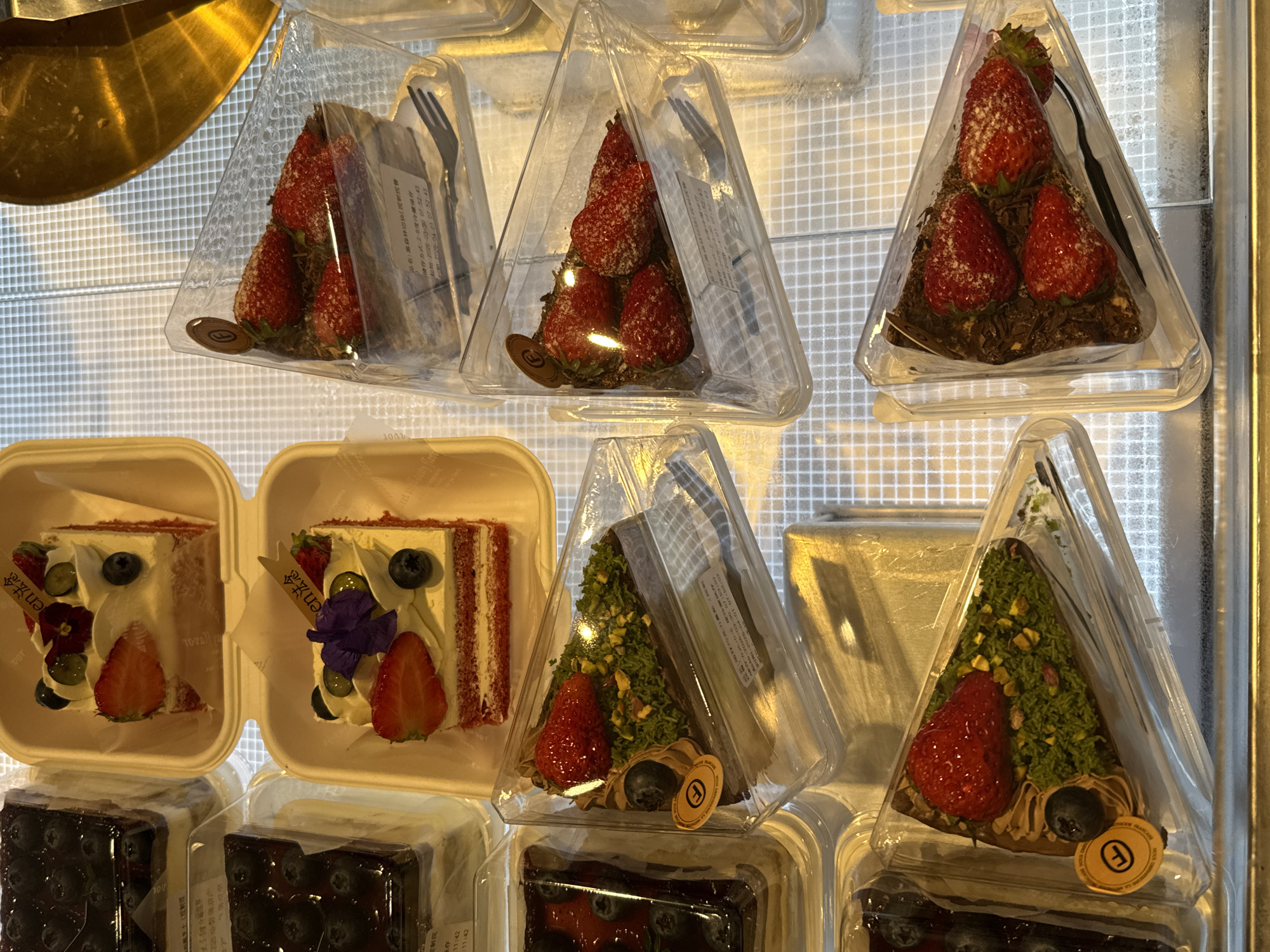 The image displays a selection of desserts displayed in a refrigerated display case. There are several types of cakes, including a red velvet cake in a square container, strawberry shortcake in clear triangular containers, and a blueberry cake in a square container. The red velvet cake is topped with cream, strawberries, blueberries, and edible flowers. The triangular containers hold the cakes with visible forks. The strawberry cakes are decorated with whole strawberries, and one variant has a green pistachio-like topping with a blueberry at the base. Each container has a label with Chinese characters and expiration dates. The display case has a grid pattern surface.