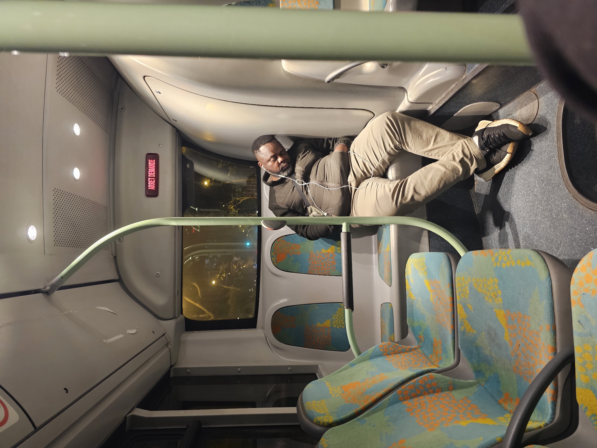 An African-American man is lying across seats on a bus, possibly sleeping. He is wearing tan pants, a dark jacket, and athletic shoes, and has white earbuds around his neck. The seats of the bus are patterned with teal, orange, and yellow designs. An "ARRET DEMANDE" sign is visible on the interior wall. Outside the window, city lights can be seen, which suggest this image was taken in the city of Paris, France, during nighttime.