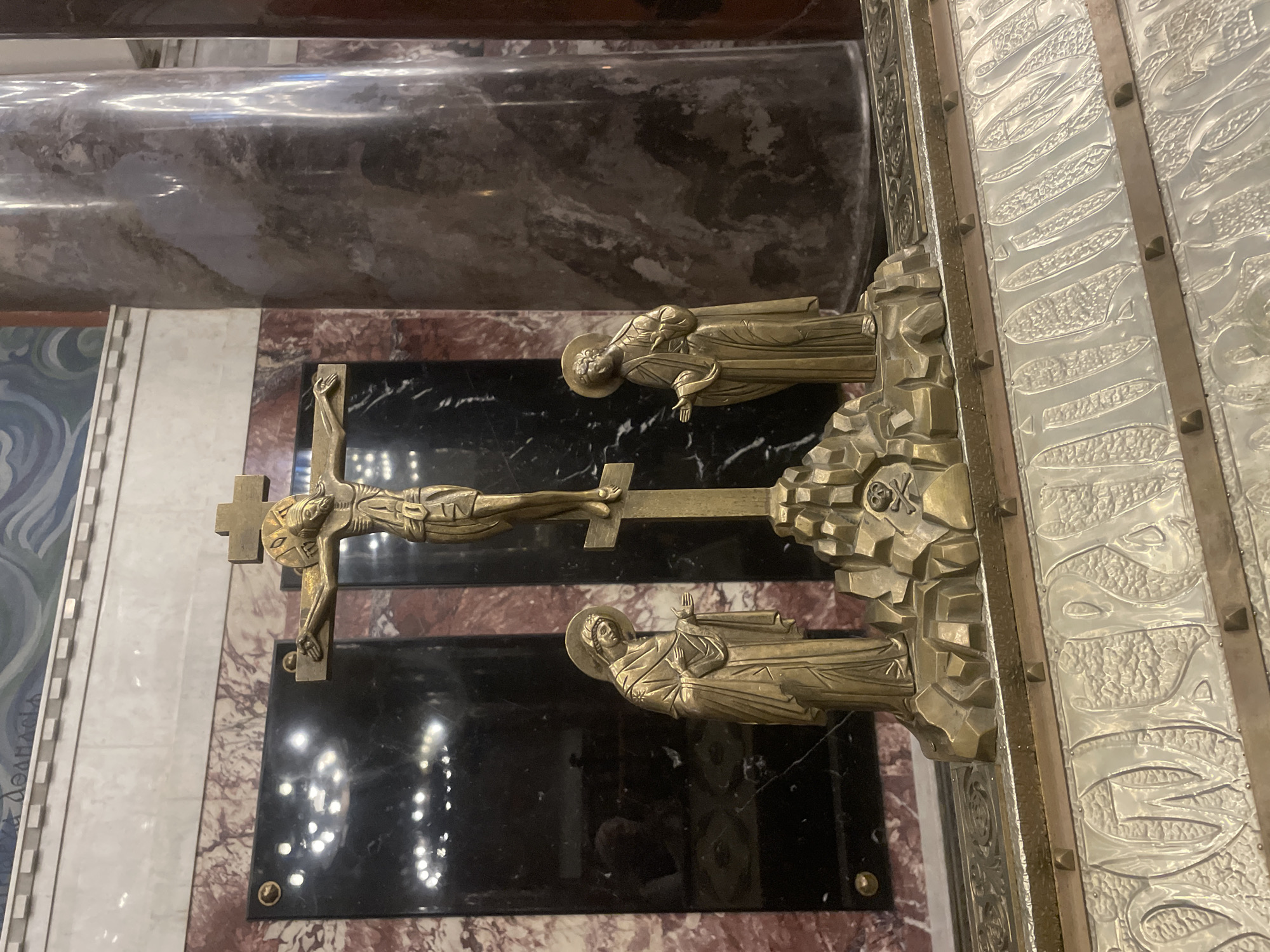 The image depicts a close-up view of a bronze sculpture of the crucifixion of Jesus Christ, placed on an altar in what appears to be a church or a religious monument. The sculpture features a central crucifix with Christ in a gilded, emaciated state. Two figures, likely the Virgin Mary and Saint John, stand on either side of the cross at its base, sculpted in a similar gilded bronze material and styled in classical robes. They are depicted with serene expressions and open hands. The base of the sculpture is rocky and textured, with a small skull and crossbones emblem visible on the right side, symbolizing Golgotha. Behind the sculpture, there are dark, reflective panels with marble bordering and a large, fluted column on the right, suggesting a grand architectural setting. The overall scene is solemn and devotional. The background features a mural with blue and white wavy patterns and a decorative cornice. Based on the style and possible context, this could be located in the Saint Nicholas Naval Cathedral in Kronstadt, Russia, a prominent landmark known for its religious significance and intricate artwork.