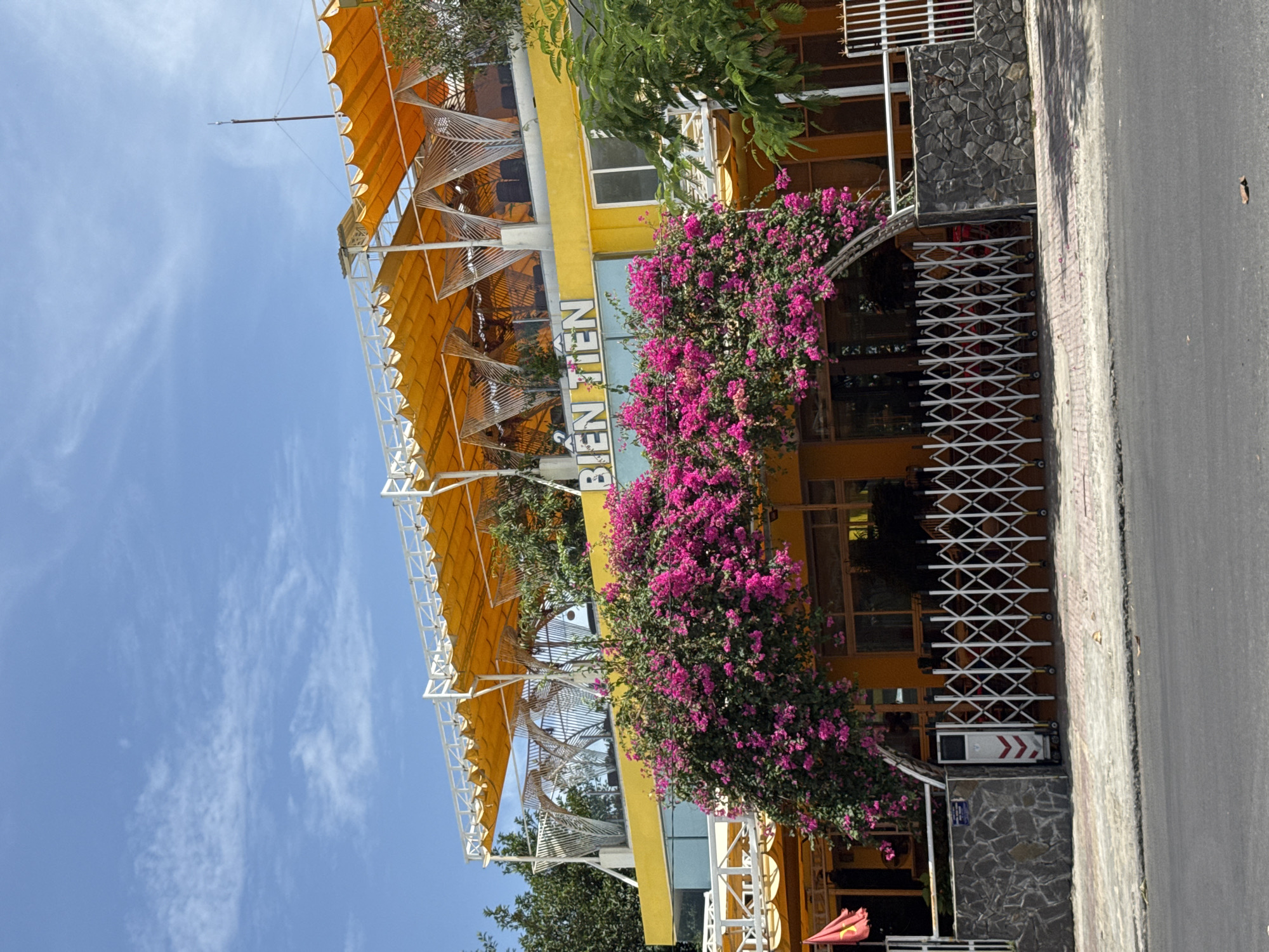 A bright yellow building with "BIEN TIEN" in blue lettering is covered in vibrant pink bougainvillea flowers. The building has multiple levels with orange awnings and white metal supports. A white retractable gate is in front of the entrance, behind a stone wall. The scene is set in Nha Trang, Vietnam, under a clear blue sky with scattered white clouds.