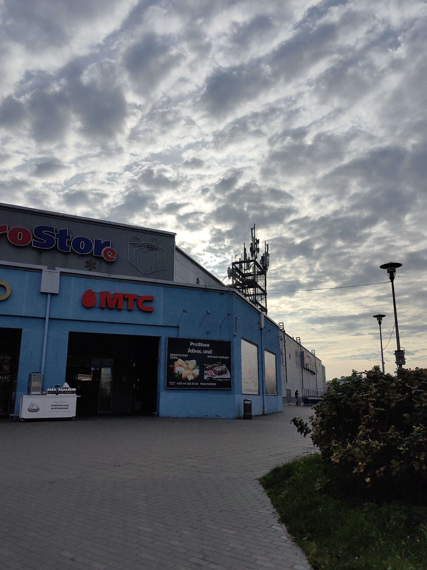 The image shows the ProStore supermarket on the outskirts of Minsk, Belarus, near the city center, at an intersection. The supermarket is a blue building with the ProStore logo and a gift icon on its facade. The MTC logo is also visible. A cell tower is located to the right of the building. The sky is cloudy and overcast. The area in front of the store is paved with interlocking concrete pavers. There are two lampposts on the right side of the image. A bush and some grass are visible in the foreground. A person is standing on the sidewalk near the building. Another person is walking away from the camera on the sidewalk further down. An ice cream cart with the text "КОБРИНСКОЕ МОРОЖЕНОЕ" is visible in front of the entrance. A poster is displayed on the wall of the building with images of cheese and meat products.