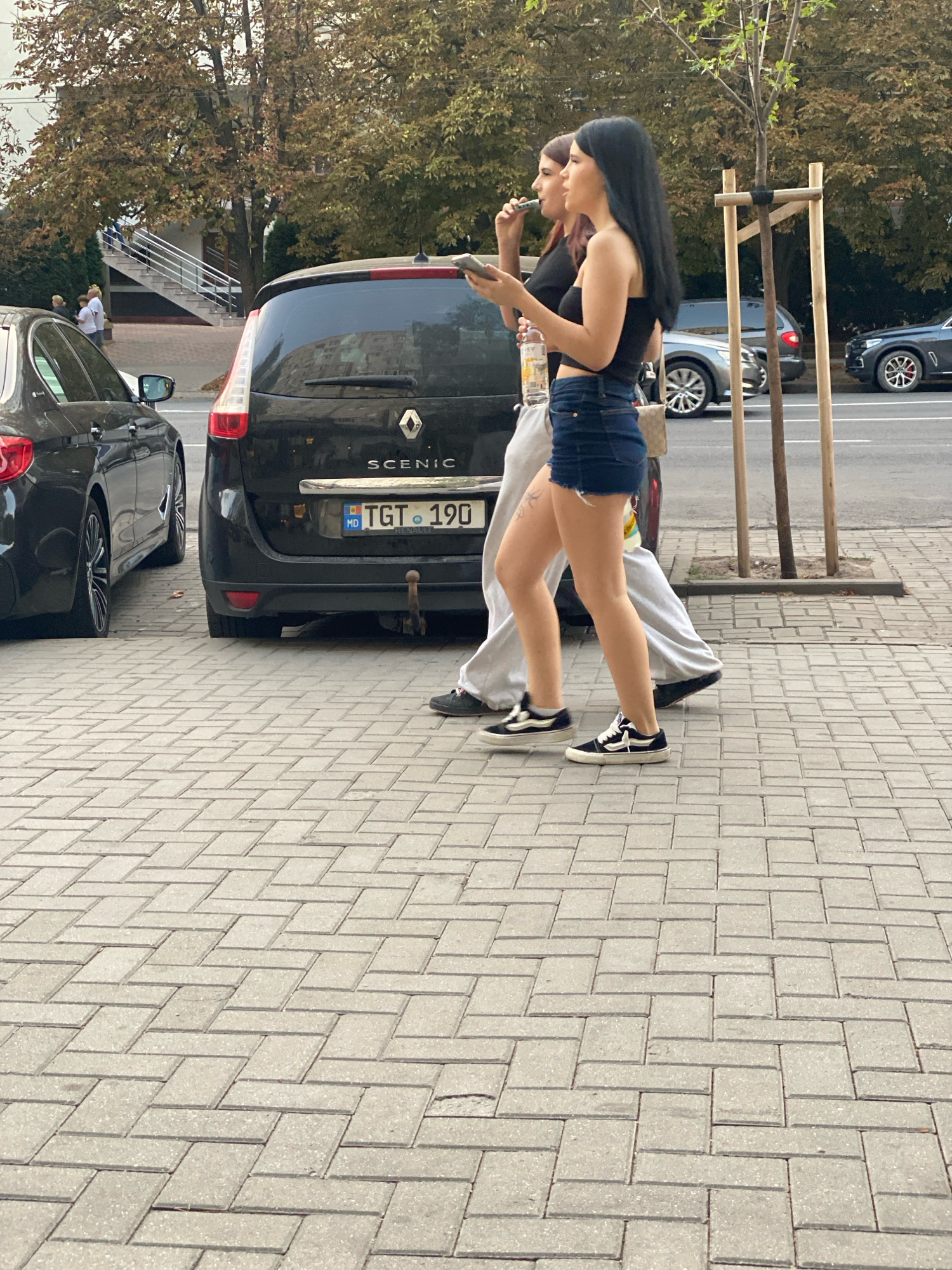 The image shows two young women walking on a paved sidewalk in Chișinău, Moldova. The woman on the right is wearing a black crop top, denim shorts, and black and white sneakers. She is holding a bottle of water in her left hand and a phone in her right. The woman on the left is wearing a black t-shirt and gray sweatpants, and appears to be smoking. Both women have dark hair.
Behind the women, several cars are parked on the street. The most prominent is a black Renault Scenic with a Moldovan license plate that reads "MD TGT 190". To the left of the Renault, a black sedan is partially visible. Further in the background, more cars and trees are visible, along with a building with a staircase leading up to it. The sidewalk is made of light-colored rectangular paving stones arranged in a herringbone pattern. The overall impression is a street scene in a city, likely during daylight hours.