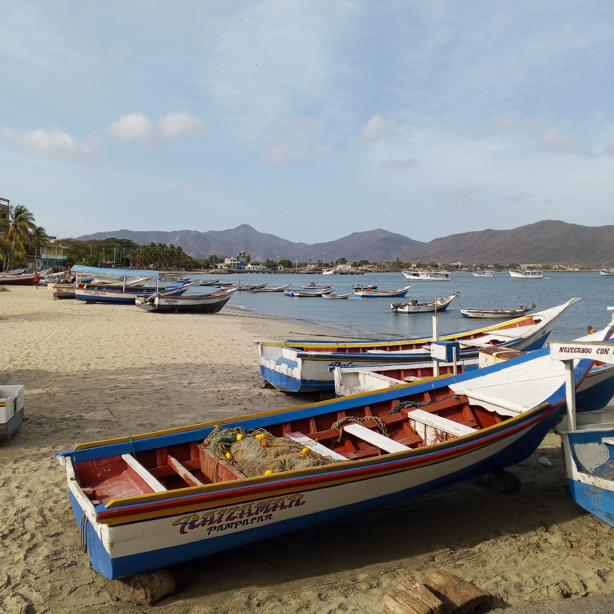 The image captures a coastal scene featuring several boats resting on a sandy beach, with a body of water visible in the background. Dominating the foreground are two fishing boats, one painted with bright colors (blue, white, red, and yellow), marked with the words "RATZAMAR. PAMPATAR," and containing fishing nets. In the distance, a line of mountains frames the horizon under a sky with scattered clouds. The location appears to be a beach, possibly in Pampatar, Venezuela, given the word on the boat. The scene is peaceful, suggesting a quiet moment along the coast.