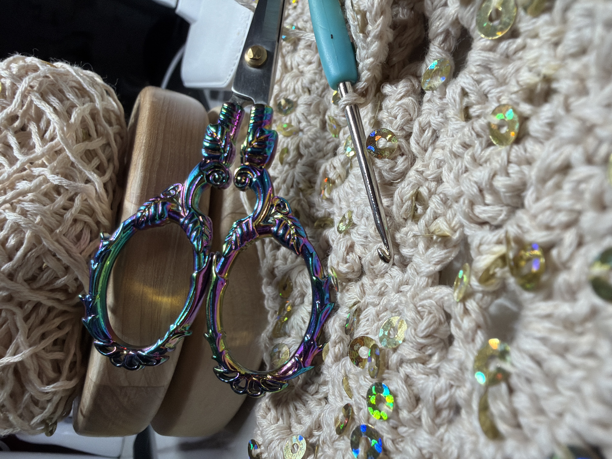 The image shows a close-up of crafting supplies. A ball of beige yarn sits to the left of a wooden object. A pair of decorative, iridescent scissors is positioned centrally. To the right, a beige crocheted fabric embellished with iridescent sequins is visible, alongside a blue-handled crochet hook. The composition is set indoors, under strong lighting conditions. There is no indication in the image that it was taken in Chaikovsky, Russia.