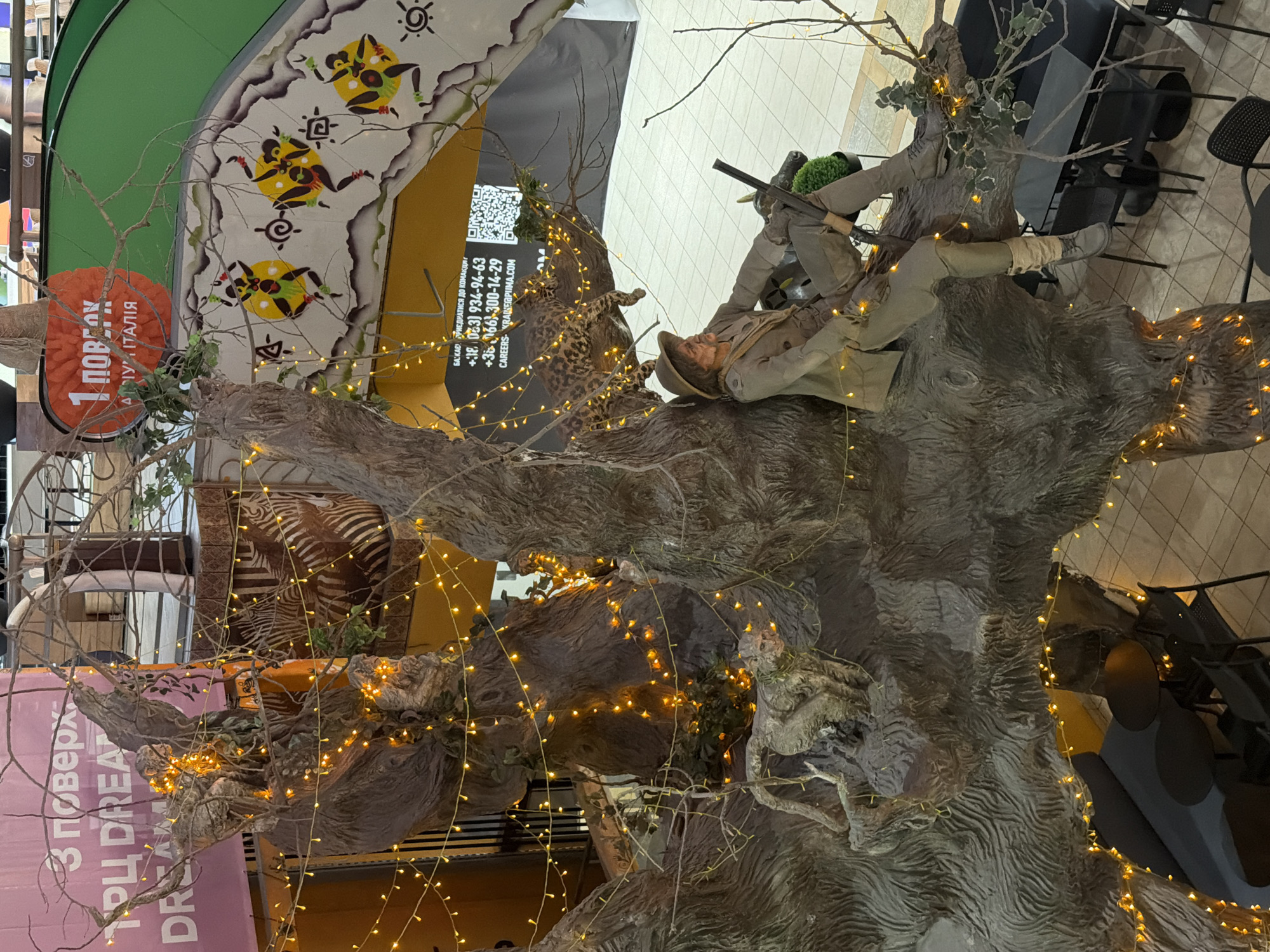 An overhead view captures a surreal display centered around a large, artificial tree structure adorned with warm, yellow fairy lights. Perched on one of the tree's thick branches is a mannequin dressed as a safari hunter, complete with a wide-brimmed hat, khaki-colored suit, and boots. The mannequin holds a rifle in its lap. Nearby, a realistic leopard sculpture is positioned on another branch. The tree itself is textured to resemble bark and appears to be part of a larger installation, possibly within a commercial or entertainment venue. In the background, various elements are visible: a large pink banner with text in Russian, an orange circular sign with Russian text, a green escalator or slide, and patterned wall art depicting stylized figures. Below the tree, cafe tables and chairs can be seen. The overall impression is one of an artistic, themed exhibit with a focus on wildlife and adventure.