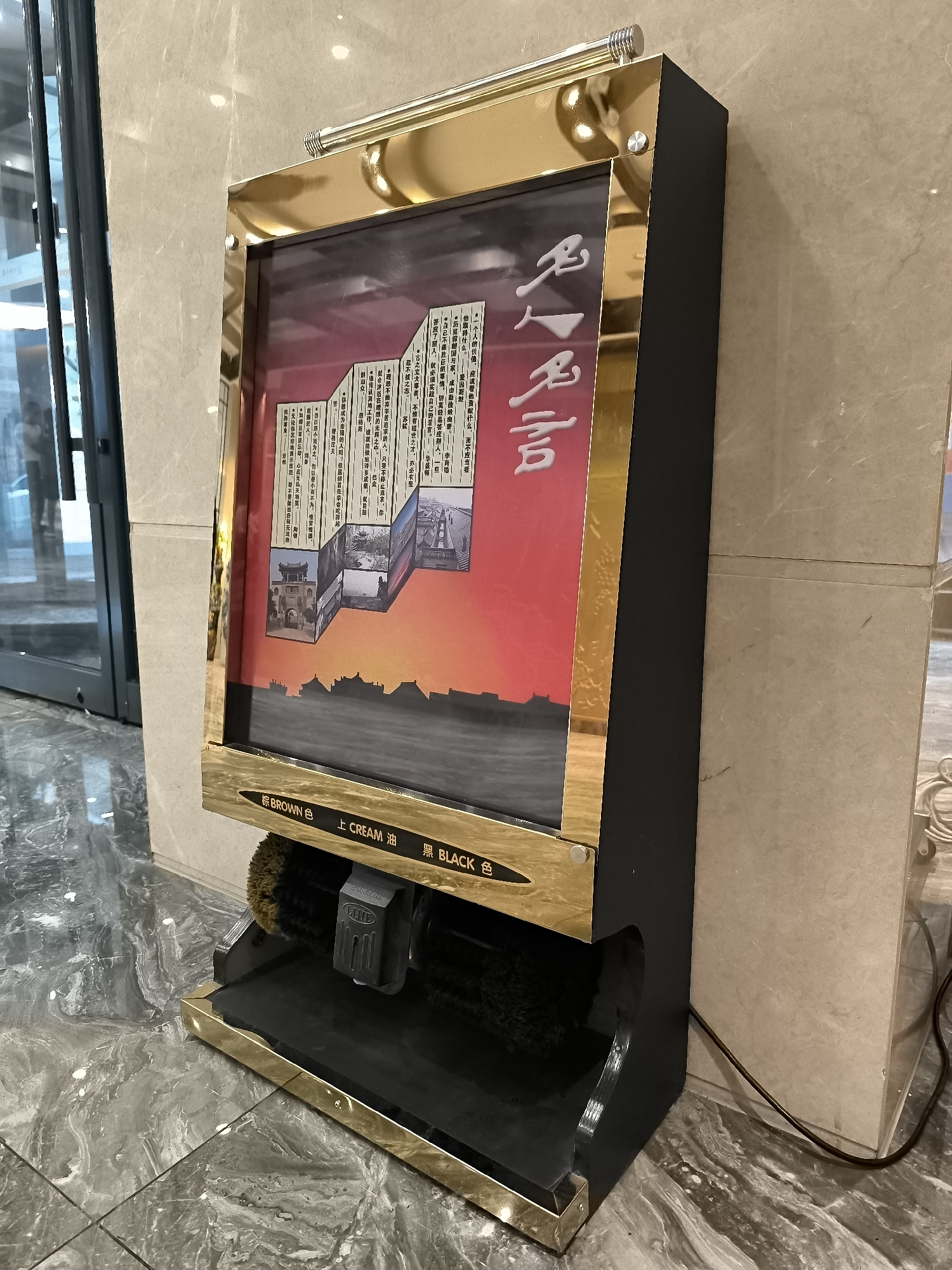 The image shows an automatic shoe shining machine located in a building's lobby in Ürümqi, China. The machine is black with gold trim and has a display screen showing an image with Chinese calligraphy and what appears to be traditional Chinese architecture. Below the screen, there are labels for "BROWN," "CREAM," and "BLACK" shoe polish, indicating the available options. The machine has rotating brushes for polishing shoes. The floor is tiled with a gray marble pattern, and the wall behind the machine is made of light-colored stone. A glass door with metal frames is visible to the left of the machine, suggesting it is situated near an entrance or exit. The lighting in the scene is bright and even, typical of an indoor commercial space. There are no people visible in the image.