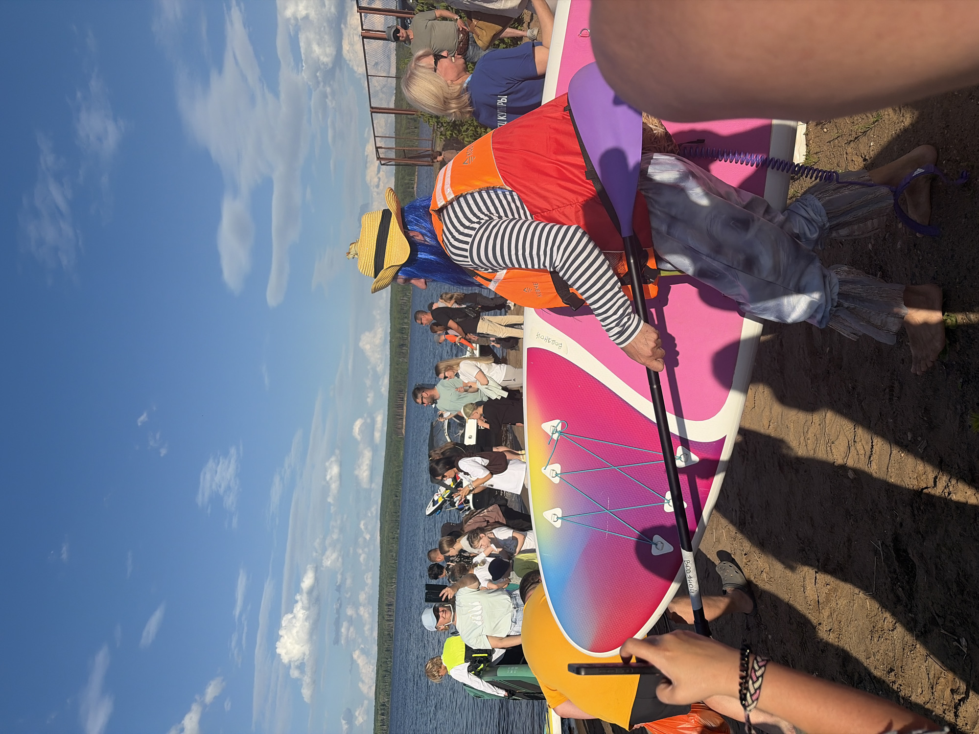 The image shows a group of people gathered on a sandy shore next to a body of water, likely a lake or river, with a clear blue sky dotted with white clouds. The scene appears to be a recreational event, possibly a paddleboarding gathering, given the prominent presence of a large, colorful paddleboard in the foreground. Several individuals are visible on and around the paddleboard, suggesting they are participating in or preparing for an activity.
One person in the foreground is dressed in a vibrant orange life vest and striped pants, with a blue wig and a straw hat, giving them a playful or costumed appearance. This individual is holding a paddle. Another person's arm with a bracelet is in the lower right corner, holding a smartphone. Other people are seen in the background, some standing on a wooden dock or pier, others sitting or interacting. The overall atmosphere seems relaxed and social, with people enjoying a sunny day outdoors. The time of day appears to be afternoon, given the bright sunlight and shadows.
There is some text visible on the paddleboard, written in a light color. One word appears to be "FondHop". Another word in what looks like Cyrillic script is partially visible on the paddle, reading "Фонд-Хоп". The location context suggests this event is taking place in Chaikovsky, Russia.