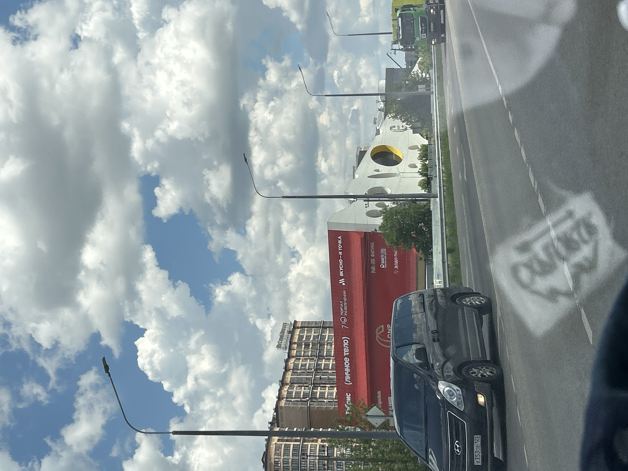 The image captures a scene from a car traveling along a road in Orlovskii, Russia. The perspective is from inside the vehicle, looking outwards. The sky is filled with large, fluffy white clouds against a bright blue backdrop, suggesting it is daytime with fair weather.
The roadside features a modern, tall building with numerous windows, possibly an apartment complex. Adjacent to it is a striking red building with prominent white lettering. Some of the visible text on the red building includes "ВКУСНО - И ТОЧКА" (Tasty - And that's it), "Табрис (личное тело)" (Tabris (personal body)), and "РИСКИЙ ФИТНЕС" (Riskiy Fitness). There's also a distinctive circular opening, a large yellow cutout, on a white structure behind the red building, adding a unique architectural element.
Several streetlights with curved poles line the road. On the road itself, the asphalt is wet, indicating recent rain or cleaning. A dark-colored minivan is visible in the foreground, its license plate reading "X3530E 123". In the distance, a green truck is also present. The overall impression is a snapshot of everyday life on a Russian road under a partly cloudy sky, with commercial and residential structures forming the backdrop.