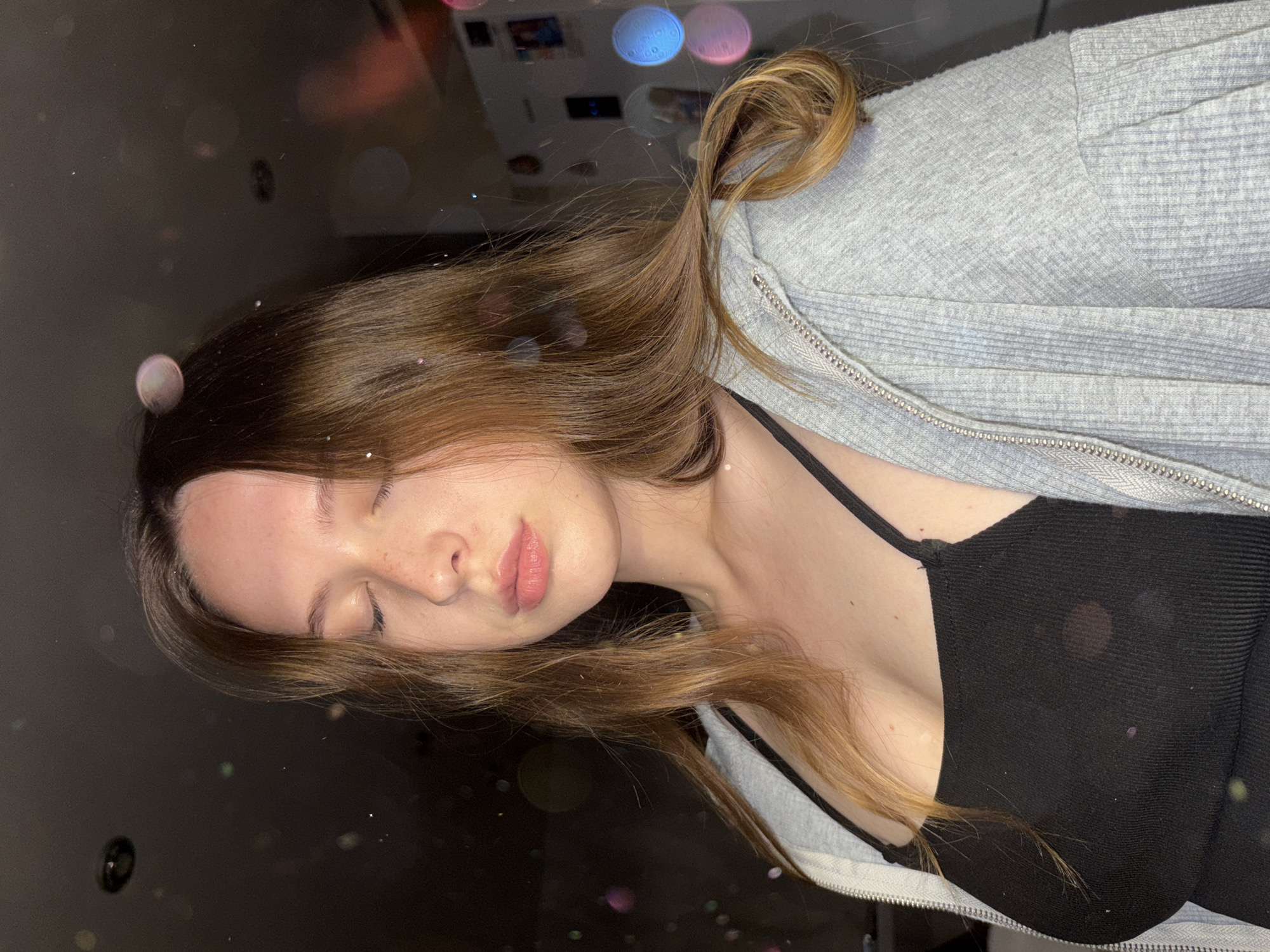 A young woman with long, light brown hair and closed eyes is pictured from the chest up. She is wearing a black tank top and a grey zip-up hoodie. The background is dark with some blurry, colorful lights that appear to be bokeh. There are also small, sparkly particles scattered throughout the image. The woman has a light dusting of freckles across her nose and cheeks. Her lips are slightly parted and appear to be glossed. The overall lighting suggests an indoor setting, possibly at night. The image does not provide any specific visual cues to indicate a location in Chaikovsky, Russia.