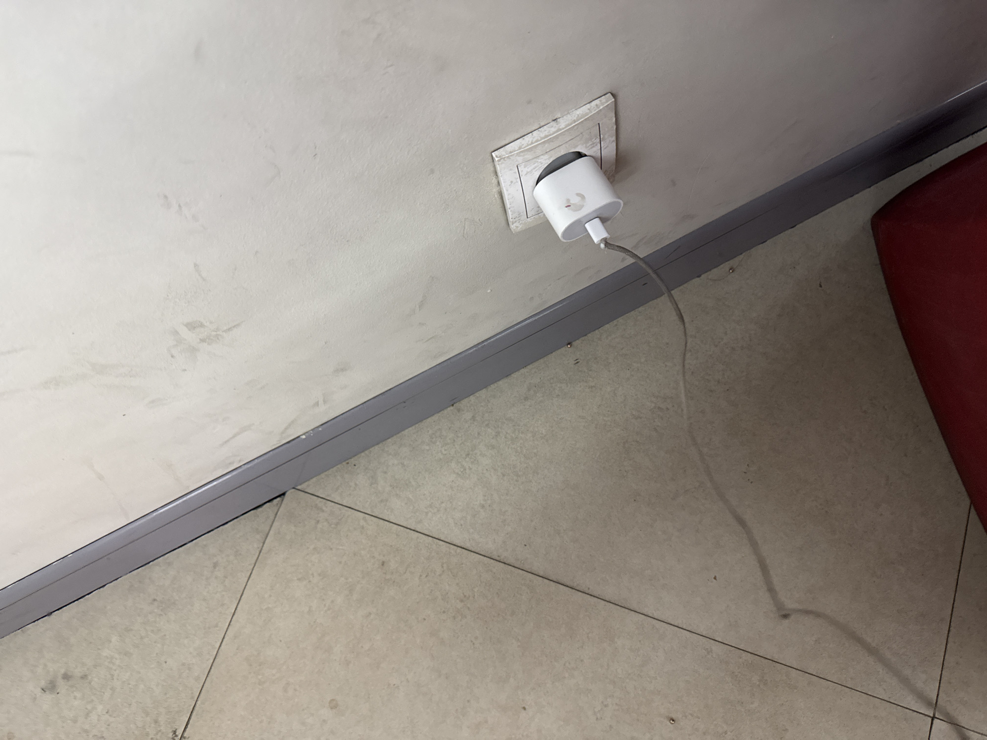 The image presents an indoor scene, likely within a building in Yerevan, Armenia, focusing on a section of a wall and floor. A light-colored, possibly off-white, wall occupies the upper and left portions of the frame, showing noticeable dirt, smudges, and stains, indicating a need for cleaning or maintenance.
An electrical outlet, recessed into the wall with a worn, off-white plastic surround, is visible towards the upper right. A white charger, which appears to be a power adapter for a portable electronic device, is plugged into this outlet. On the side of the charger, a faint, unidentifiable logo or symbol, possibly resembling a stylized bird or animal, is discernible. A light-colored cable extends downwards from the charger, draped onto the floor.
A grey skirting board runs horizontally along the base of the wall, separating it from the floor. The floor is tiled with light-toned, possibly beige or pale grey, square tiles, which also exhibit signs of dirt and wear, particularly along the grout lines and surfaces. In the bottom right corner, a portion of a red object, likely a piece of furniture such as a chair or sofa, is visible, adding a contrasting color element to the otherwise neutral and somewhat drab environment. No people are present, and the lighting suggests an indoor setting with no clear indication of the time of day.