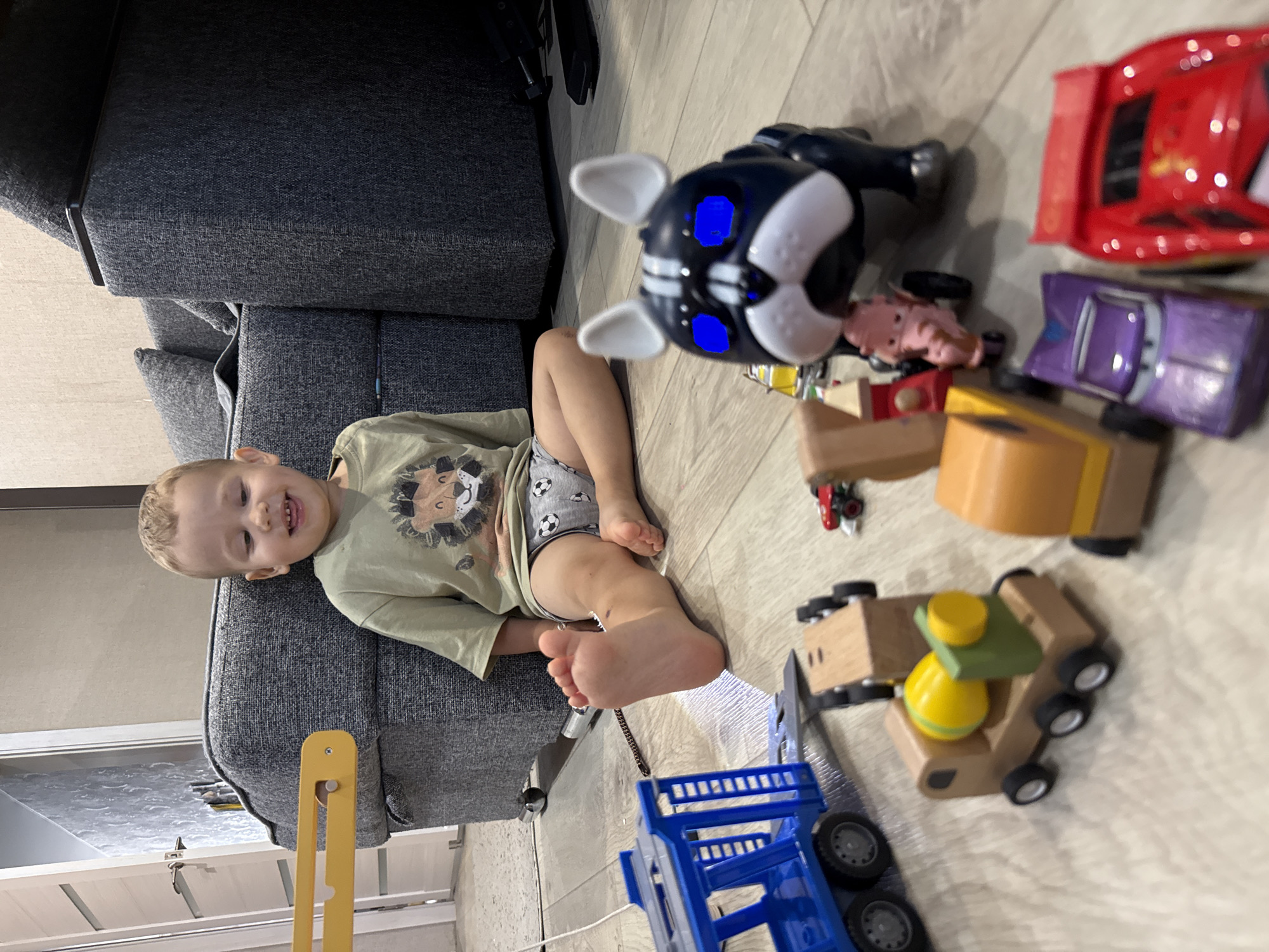 A young boy with blond hair is sitting on a light wood floor in what appears to be a home in Rusanovo, Russia. He is wearing a light green t-shirt with a lion graphic and grey shorts with a soccer ball pattern. The boy is smiling and looking to his left. He has a bright blue toy dog with white accents and illuminated blue eyes in front of him. Scattered around the boy are various toys, including a blue toy truck, a wooden cement mixer, a wooden train car, a small red toy car, a pink toy pig, a purple toy car, and a red toy race car. A grey couch is visible behind the boy, and a doorframe with a white door is to his left. The environment appears to be a living room.