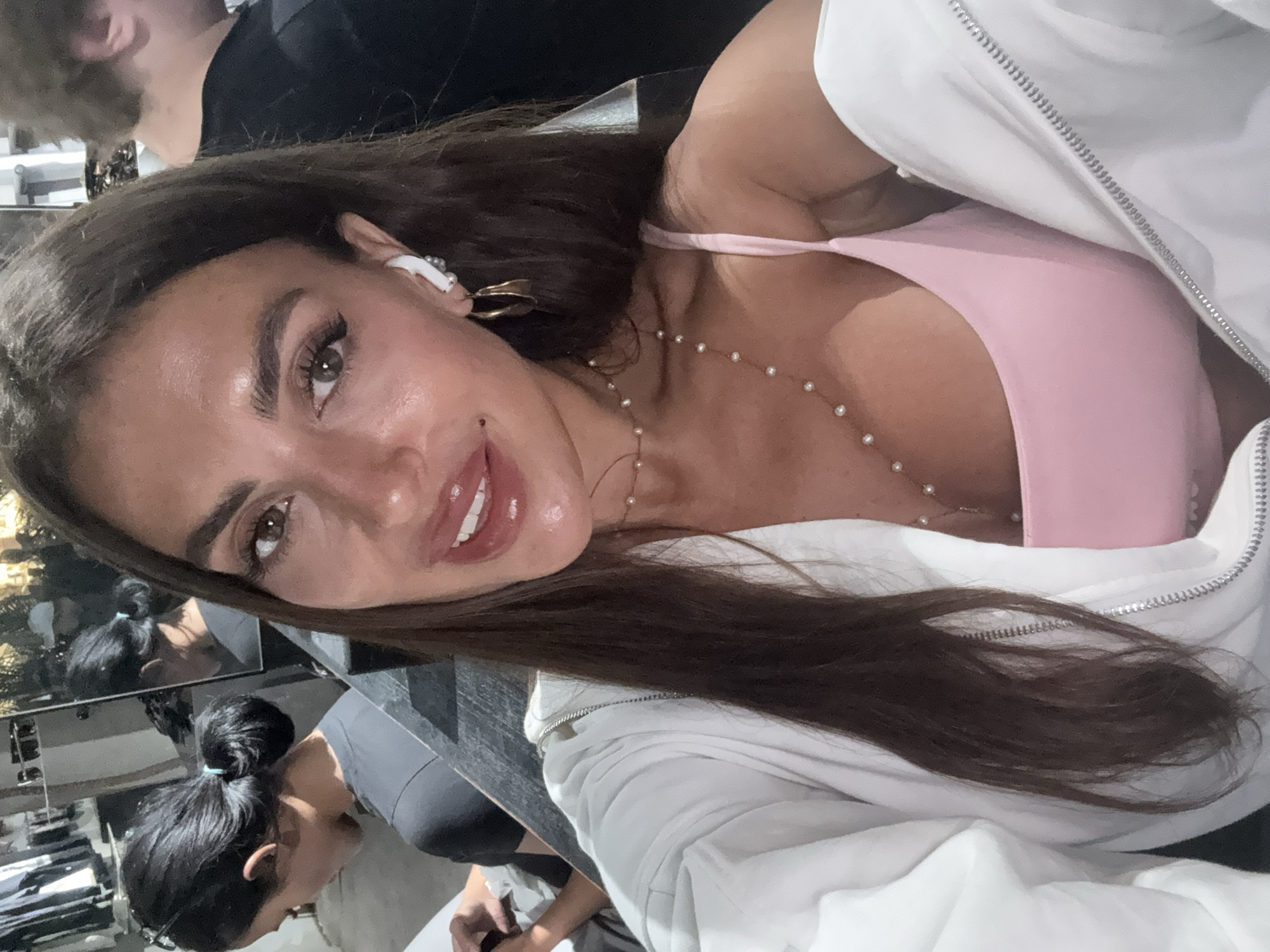 A woman with long brown hair is wearing a pink top and white jacket, along with a pearl necklace. She has light skin, brown eyes, and is wearing an earring and an earpiece. The location is North Kuta, Indonesia. The image includes a reflection of another woman with dark hair in a bun, standing near a rack of clothes. There is a man with short hair in the background.