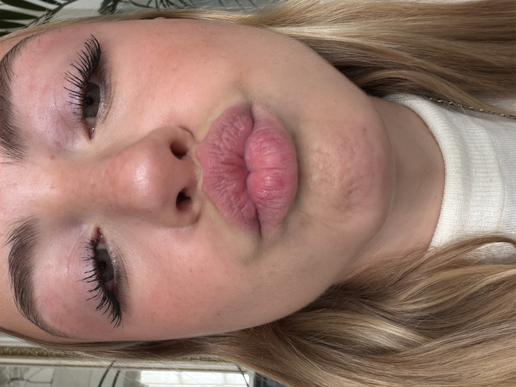 The image presents an upward-angled, close-up portrait of a young adult female, depicted from the mid-chest upwards. Her light-toned skin exhibits some light freckling on the nose and cheeks. Her eyes feature dark irises and prominently mascara-enhanced eyelashes. Her lips are noticeably pursed into a puckering or kissing gesture. Light blonde hair cascades around her face and shoulders. She is dressed in a white ribbed turtleneck sweater, with a faint chain necklace visible near the collarbone. The background is blurred and indistinct, indicating an indoor setting, with visual cues including a partial, ornate golden frame on the left and blurred green foliage, consistent with a houseplant, on the right. The specific location in the city of Paris, France, cannot be determined from the visual evidence.