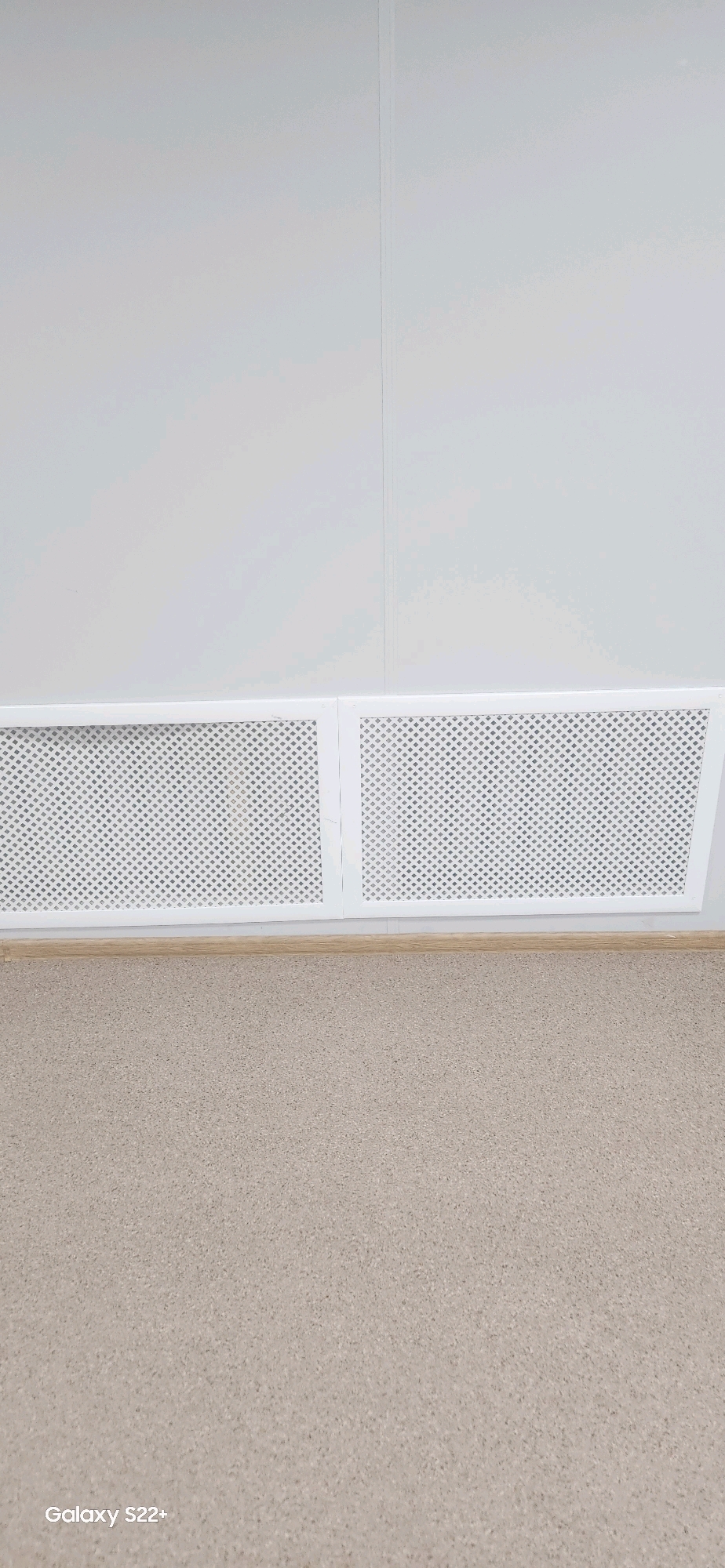The image shows a section of a room with a white wall and a light-colored, textured floor. Along the bottom of the wall, there are two white decorative grilles, possibly for a heating system or ventilation. A light brown baseboard runs between the grilles and the floor. The floor appears to be carpeted or covered with a similar material. In the bottom left corner of the image, the text "Galaxy S22+" is visible, indicating it is a photo taken with a Samsung Galaxy S22+ phone. The scene is well-lit and appears to be indoors, with no windows or doors immediately visible. There are no people or animals present in the image. The overall impression is one of a clean, minimalist interior. The image does not contain enough information to determine the specific location within the city of Nyurba, Russia, other than it is an indoor scene.