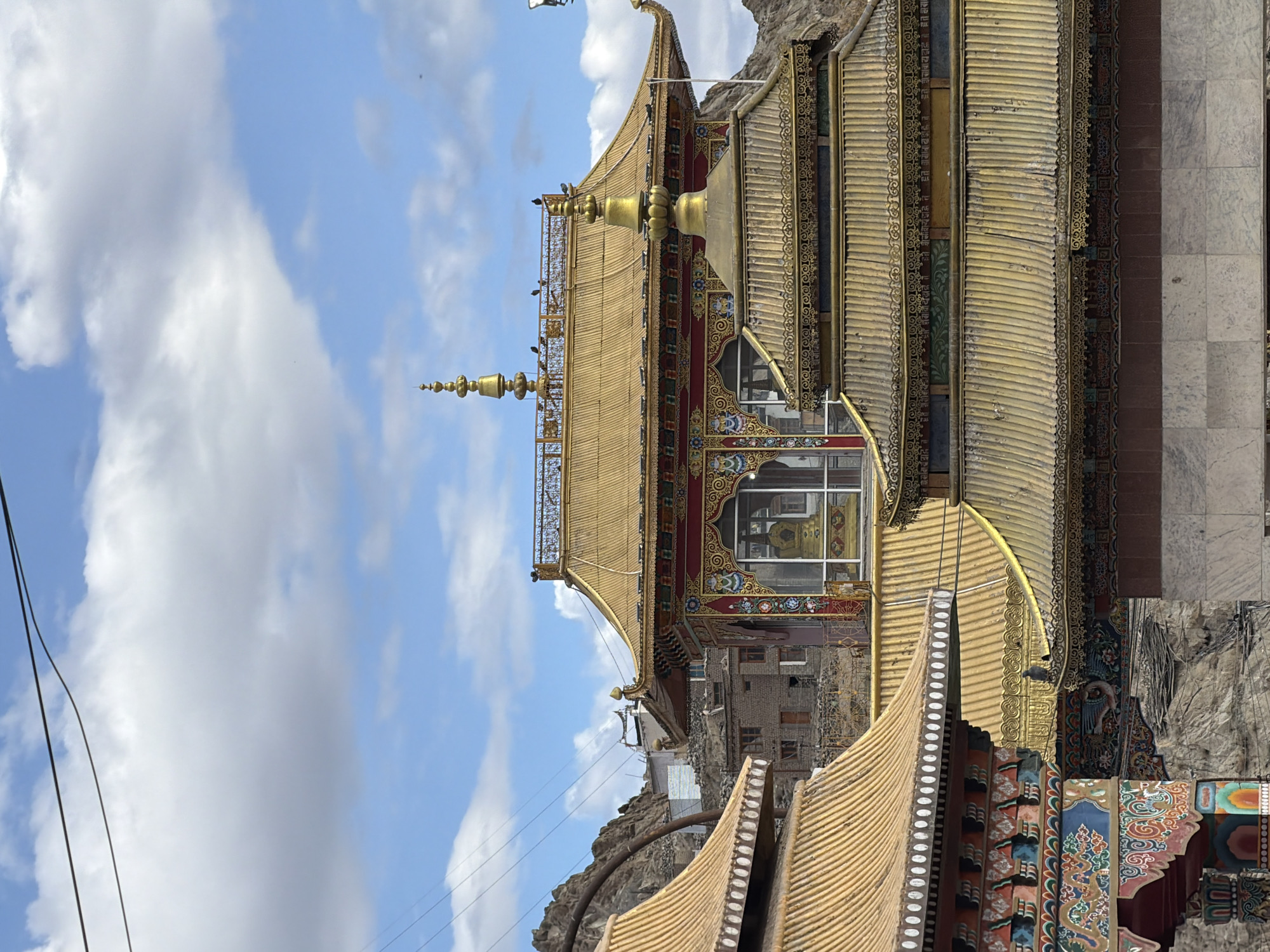 The image shows the golden roofs of a monastery in Leh, India, against a cloudy blue sky. The architecture is distinctly Tibetan Buddhist, with tiered roofs adorned with intricate golden ornamentation and prayer flags. In the foreground, a colorful Buddhist temple structure with painted motifs is visible on the left. Behind it, other golden rooftops and parts of the monastery complex are seen, including a building with a large golden stupa inside a glass enclosure. The background features the rugged, rocky terrain characteristic of the Leh region, with some traditional Ladakhi buildings nestled into the hillside. Power lines are visible stretching across the sky, indicating the presence of modern infrastructure in this otherwise traditional setting. The overall scene conveys a sense of religious and cultural heritage within a mountainous landscape.